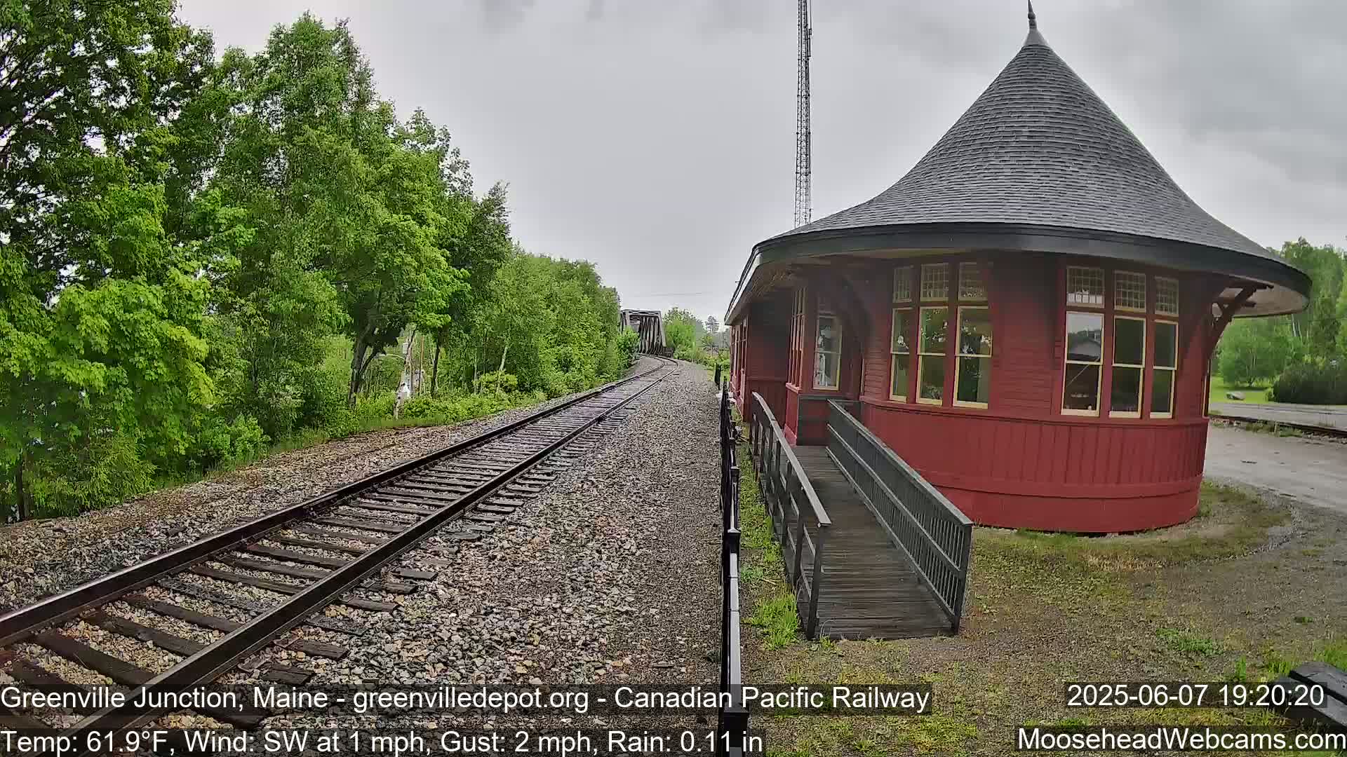 A red, round train depot with a ramp sits beside railroad tracks that curve into the distance under a gray, rainy sky, surrounded by lush green trees.