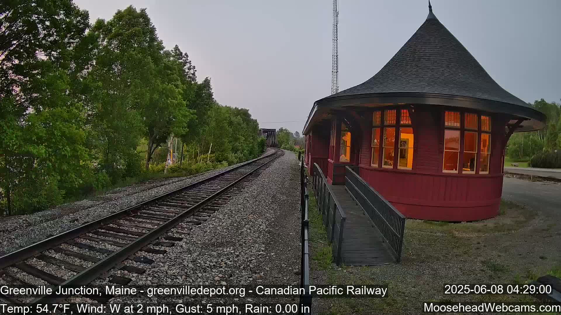 A red, round train depot with lit windows sits beside railroad tracks that wind through a wooded area under a mostly clear, dusky sky.