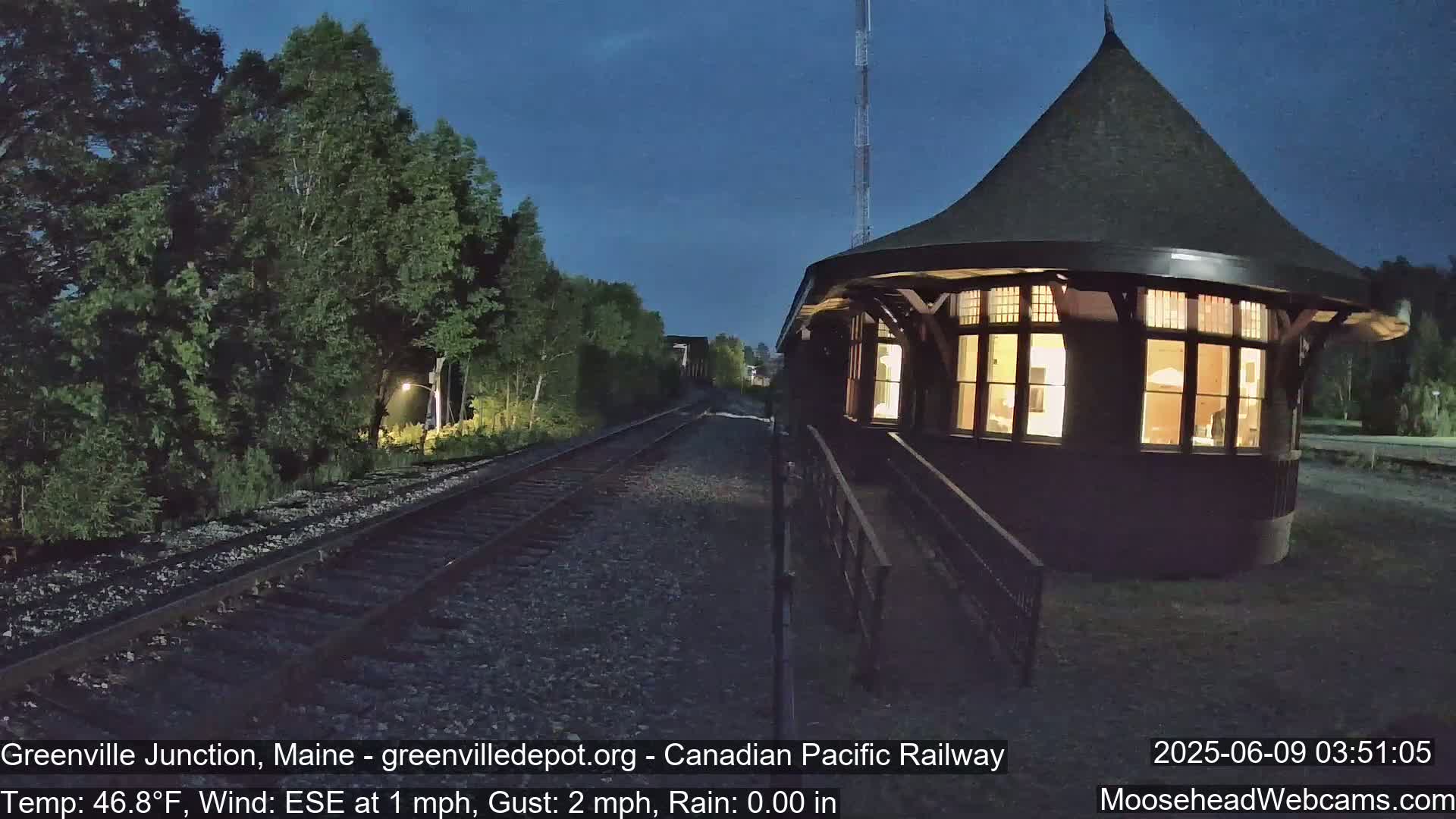 A lighted, round train depot stands beside railroad tracks at night under a dark blue sky.