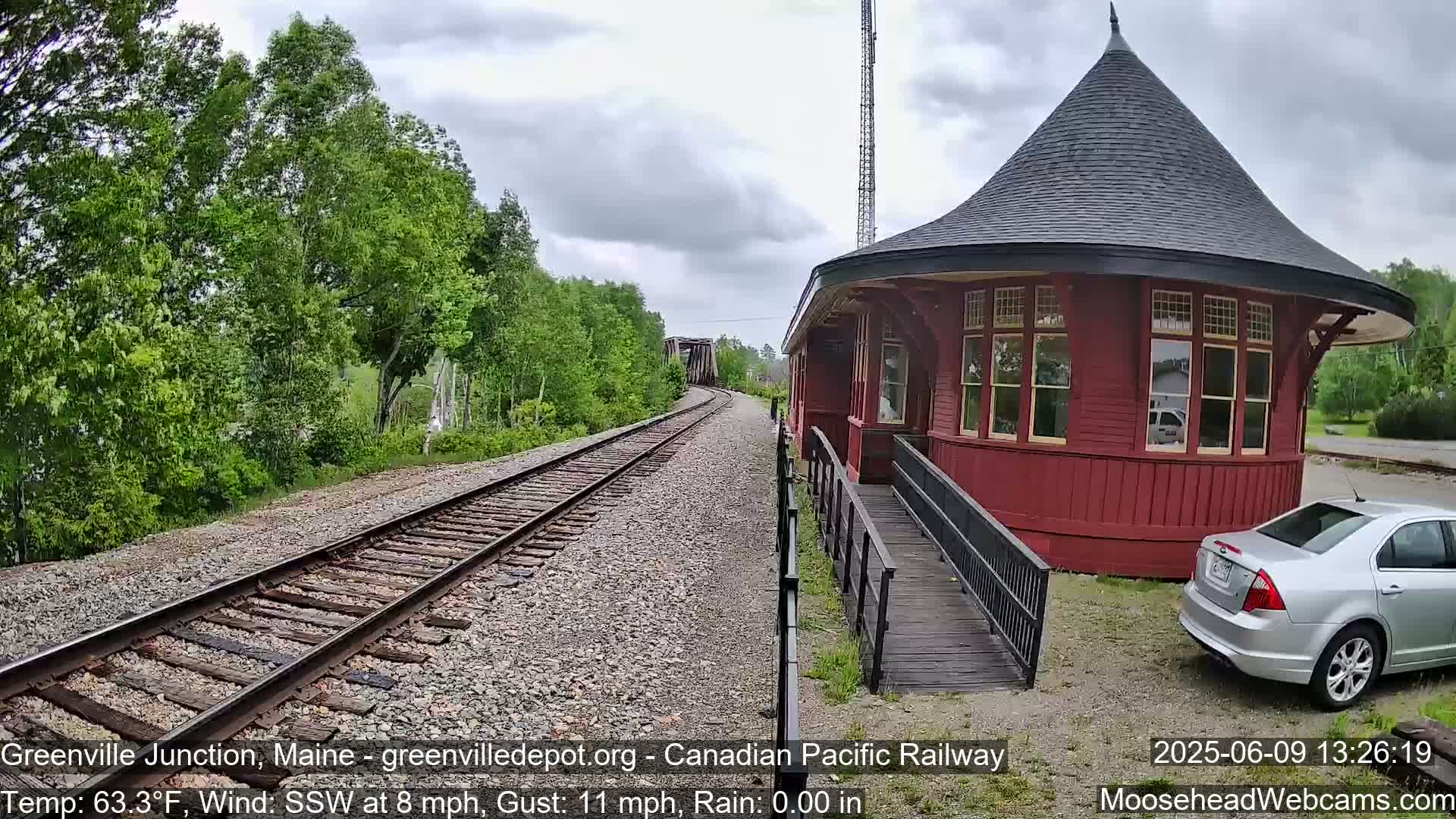 A red, round train station with a silver car parked nearby sits beside train tracks curving away into a wooded area under a cloudy sky.
