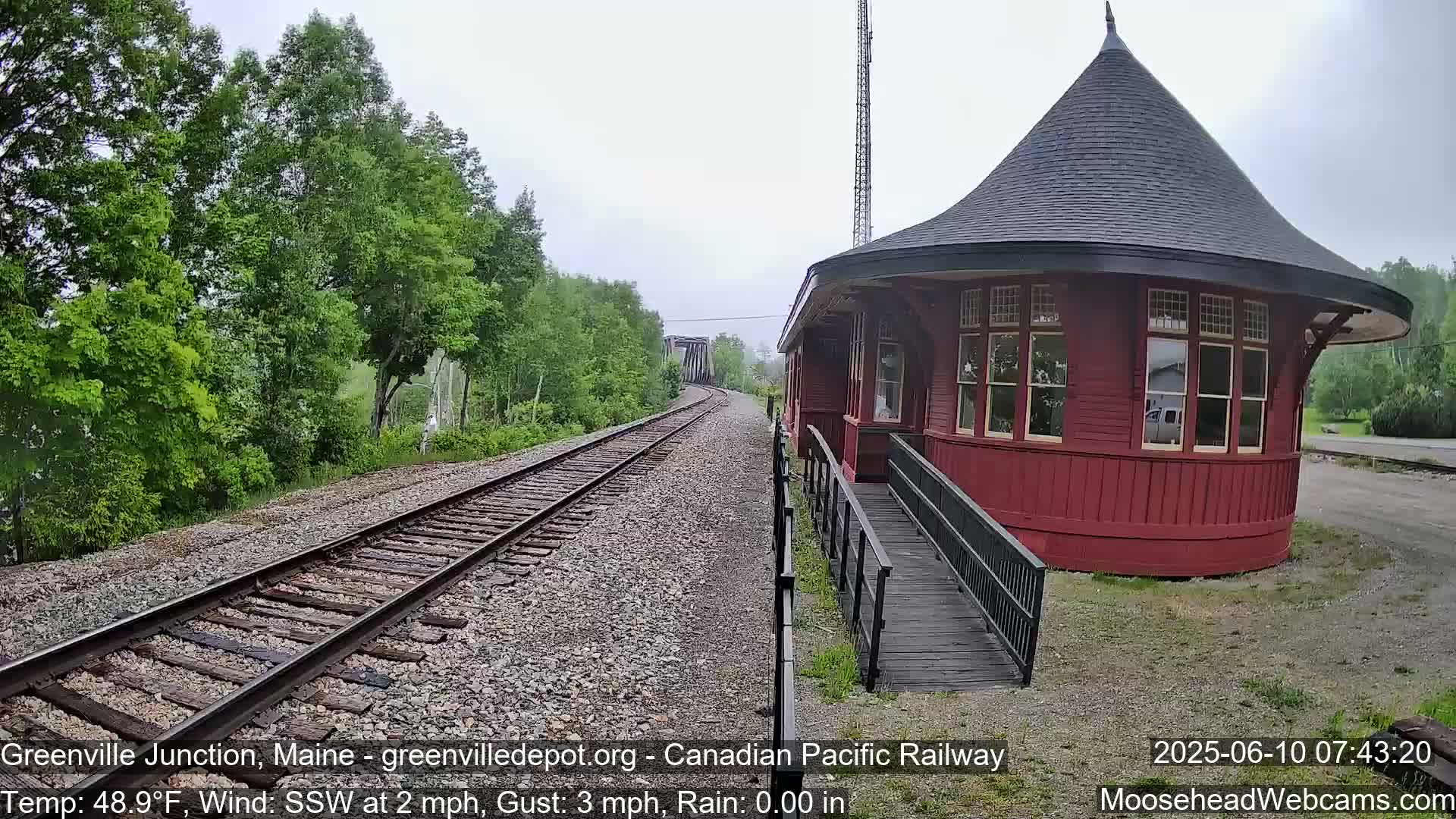 A red, round train depot with a ramp sits beside railroad tracks that curve into the distance under a cloudy sky, surrounded by lush green trees.
