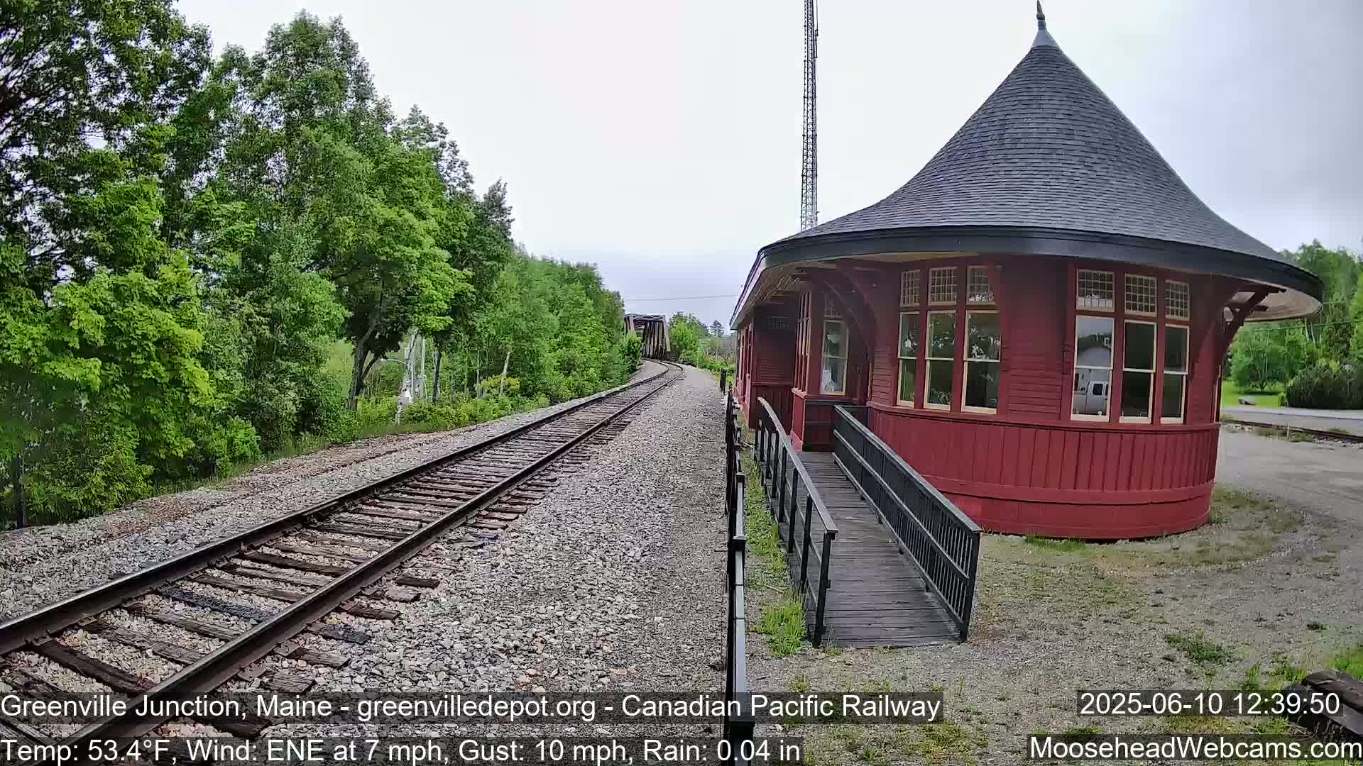 A red, round train station with a ramp sits beside railroad tracks that curve into a lush green forest under a light rain.