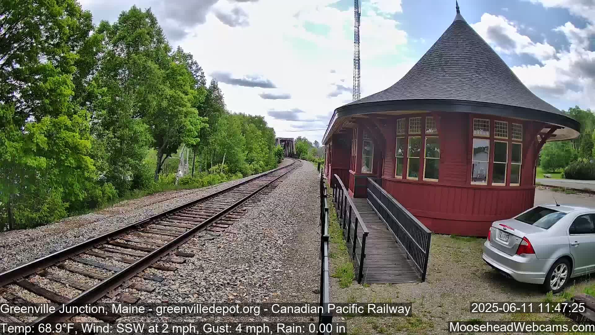 A red train station with a conical roof and a ramp sits beside railroad tracks that extend into a lush green landscape under partly cloudy skies with light rain.