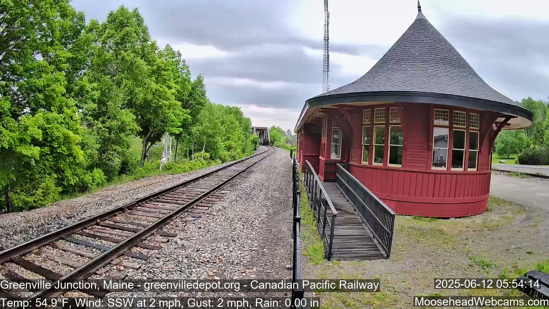 A red, round train depot with a conical roof stands beside railroad tracks on a rainy day, surrounded by lush green trees.