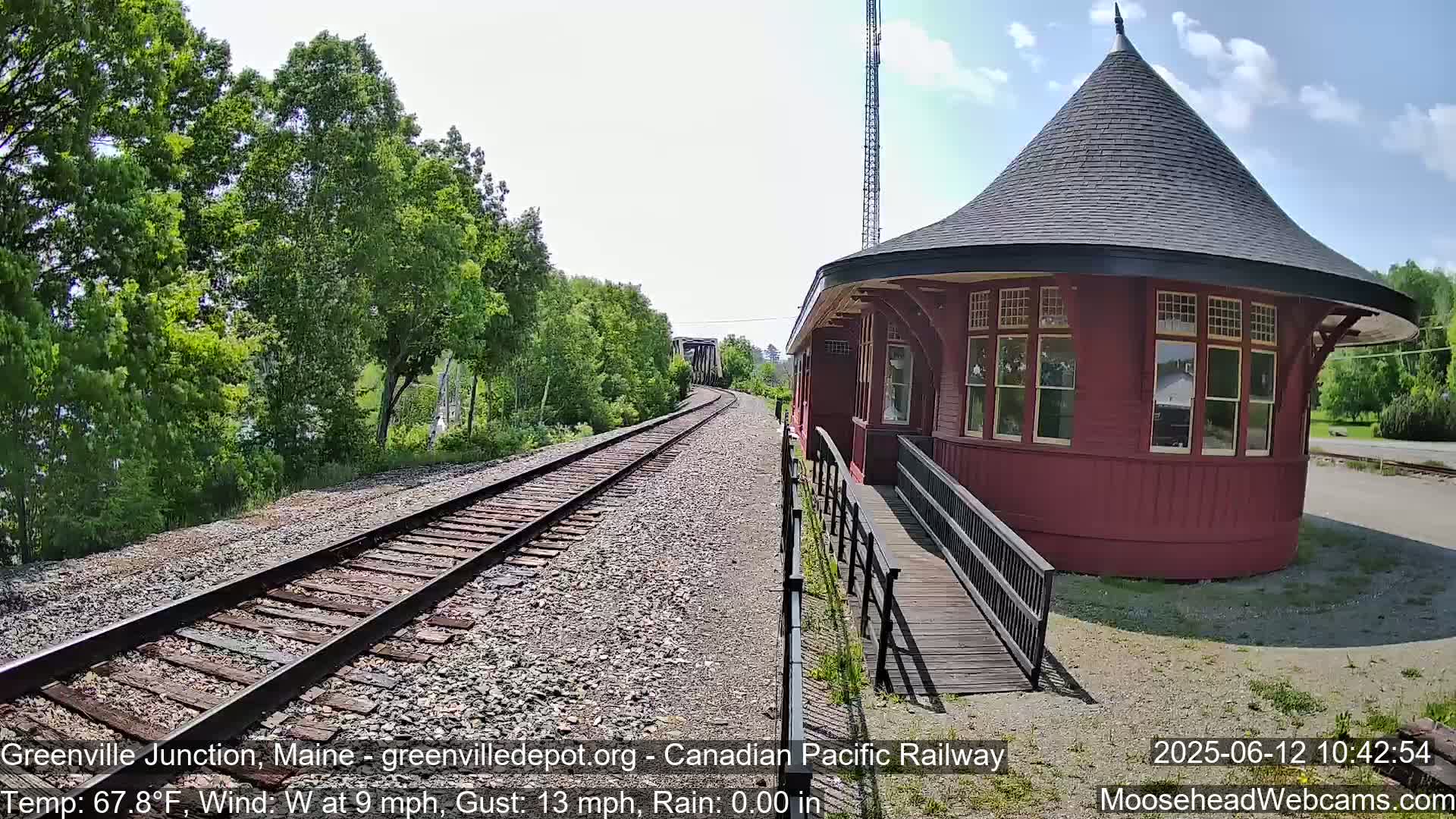 A red, round train depot with a conical roof sits beside train tracks that curve gently into the distance, under a partly sunny sky.