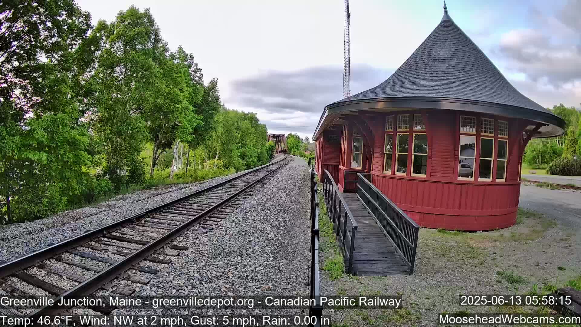 A red, round train depot with a conical roof sits beside train tracks that disappear into a green, wooded area under a partly cloudy sky.