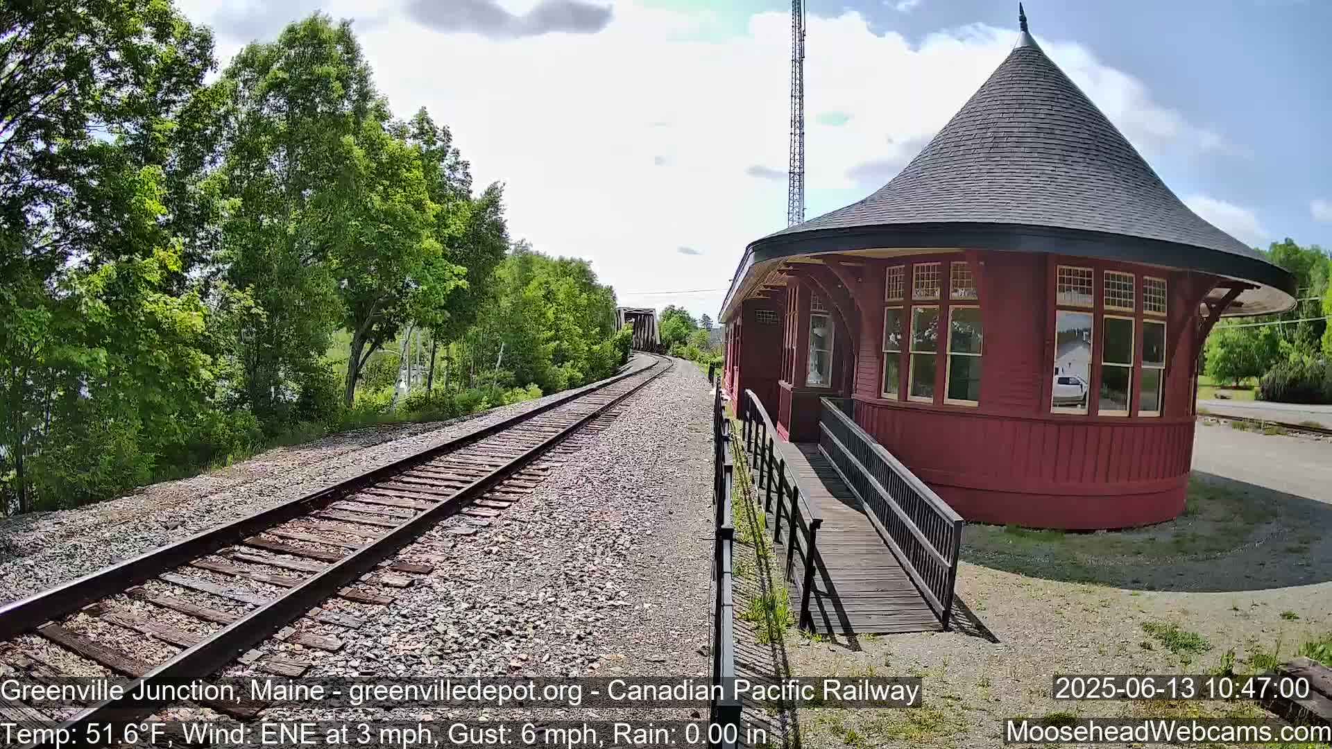A red, round train depot with a peaked roof sits beside railway tracks that extend into a landscape of green trees under a partly cloudy sky.