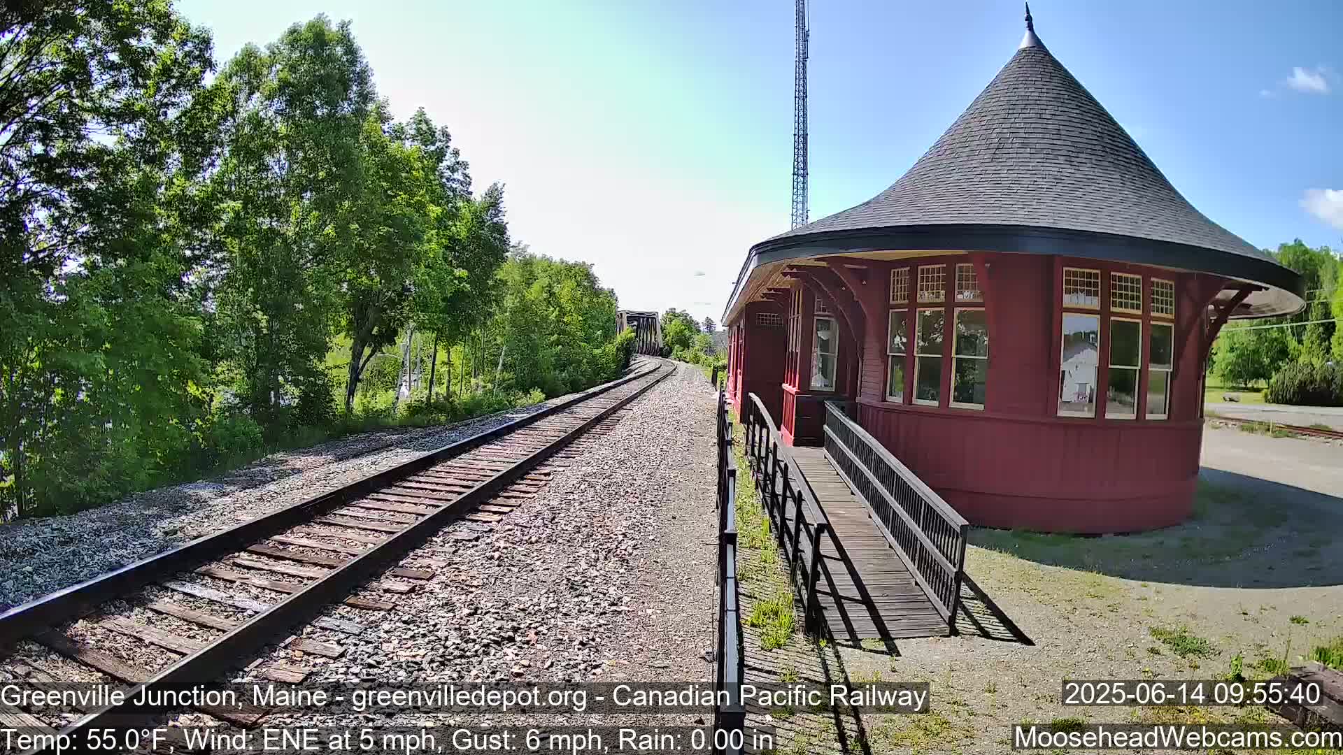 A red, round train depot with a ramp stands beside railroad tracks on a sunny day with a light east-northeast wind.