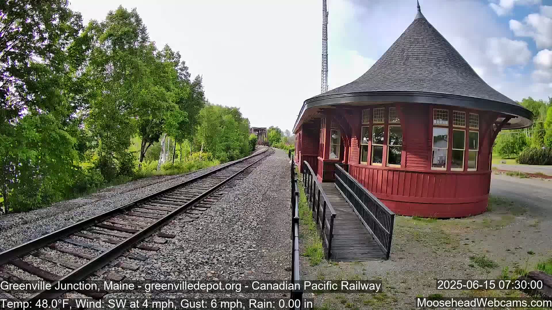 A red, round train depot with a conical roof sits beside railroad tracks on a partly sunny day with a southwest wind.
