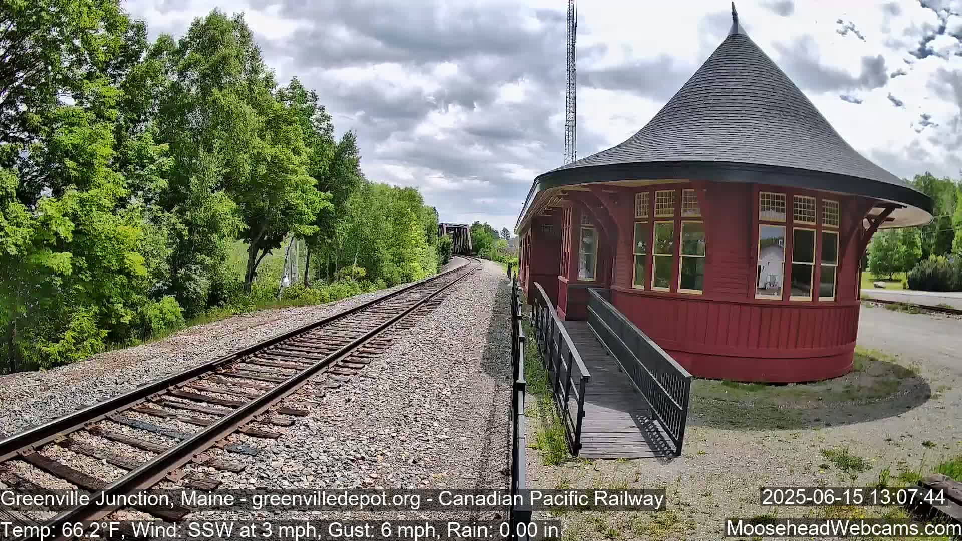 A red, round train station with a conical roof sits beside railway tracks that extend into a lush green forest under a partly cloudy sky, with light rain.