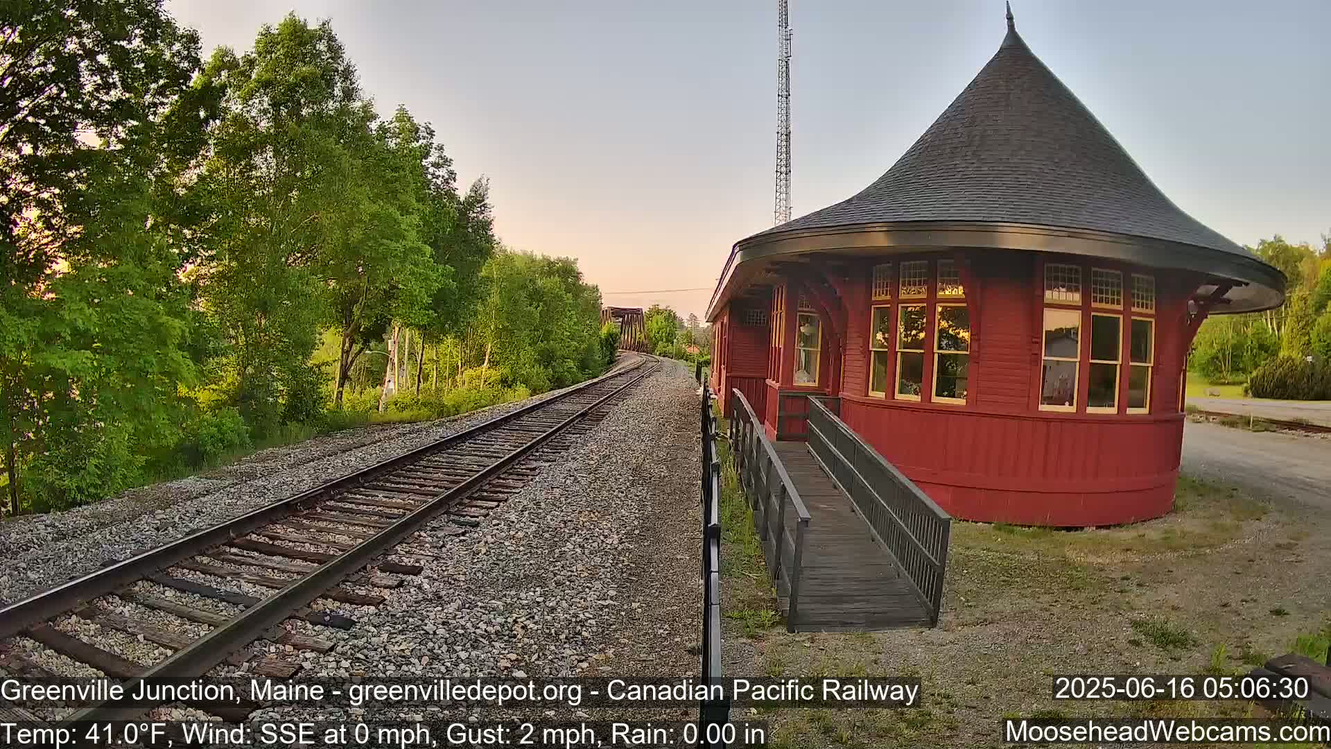 A red, round train depot with a conical roof stands beside railroad tracks leading into a wooded area under a light, partly cloudy sky.