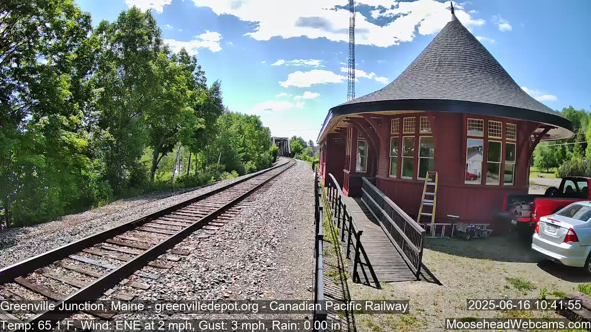 A red train depot with a conical roof sits beside railroad tracks on a partly sunny day with a light east-northeast wind.