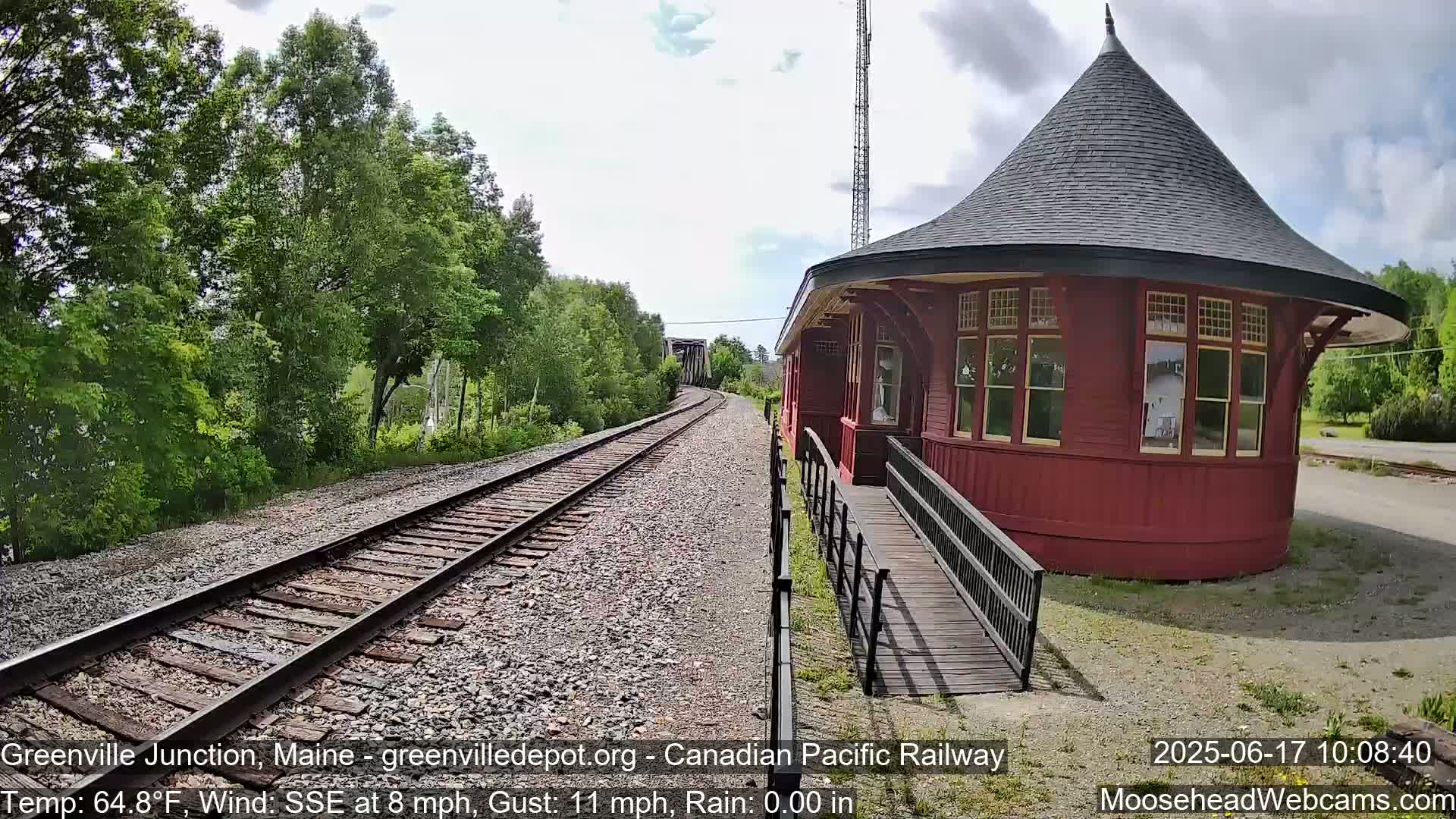 A red, round train depot with a conical roof stands beside railroad tracks leading to a bridge under a partly cloudy sky, with a light wind and no rain.