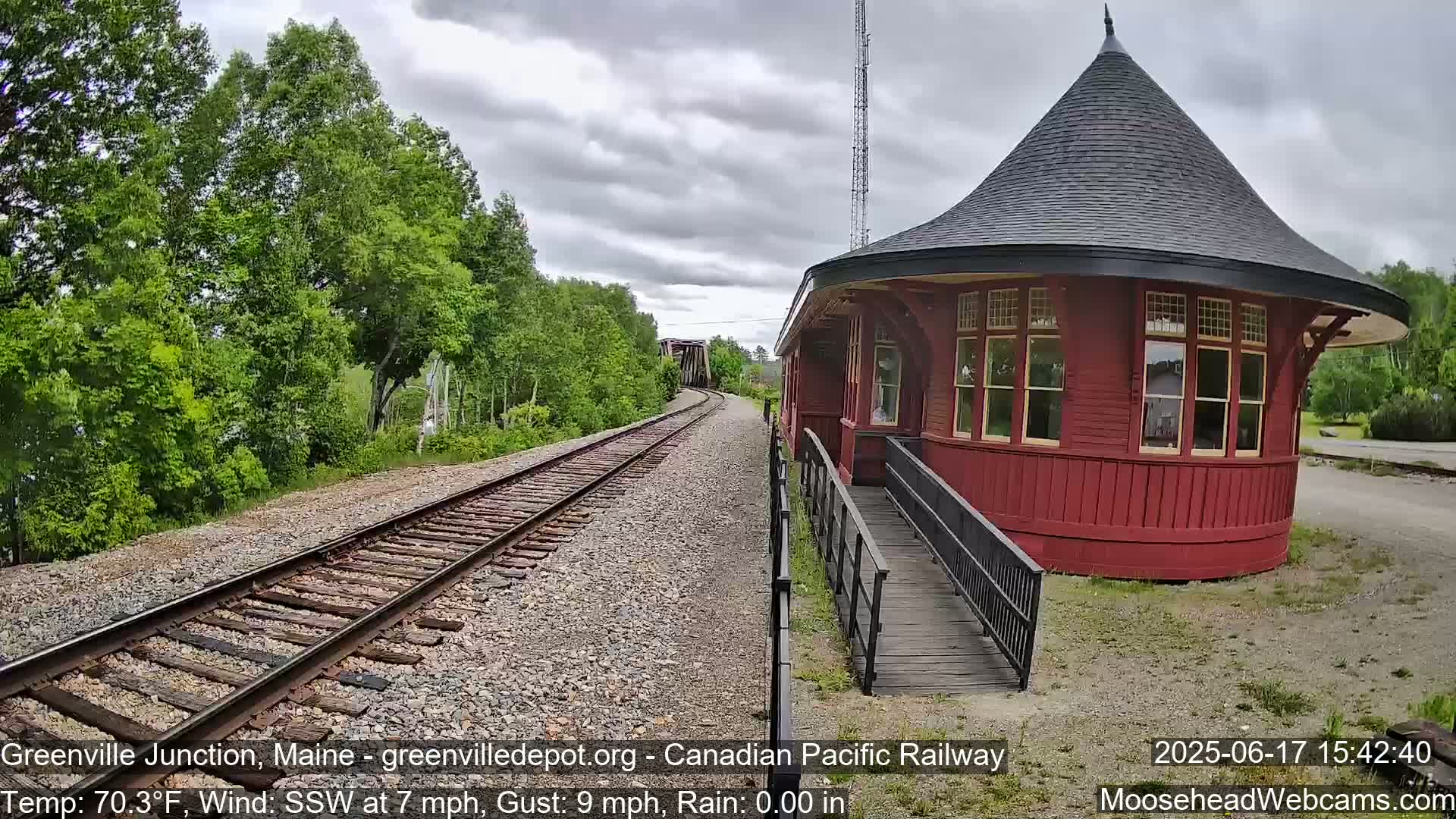 A red, round train depot with a ramp is situated beside railroad tracks that extend into the distance under a cloudy sky with light rain.