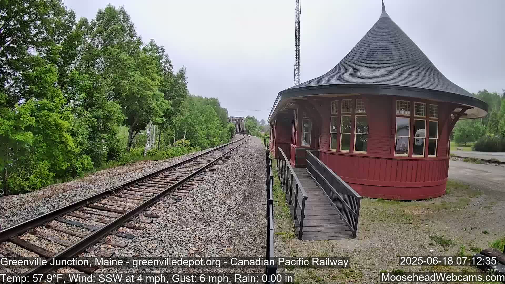 A red, octagonal train station with a conical roof sits beside railroad tracks that curve into the distance, surrounded by green trees under a cloudy sky with light rain.