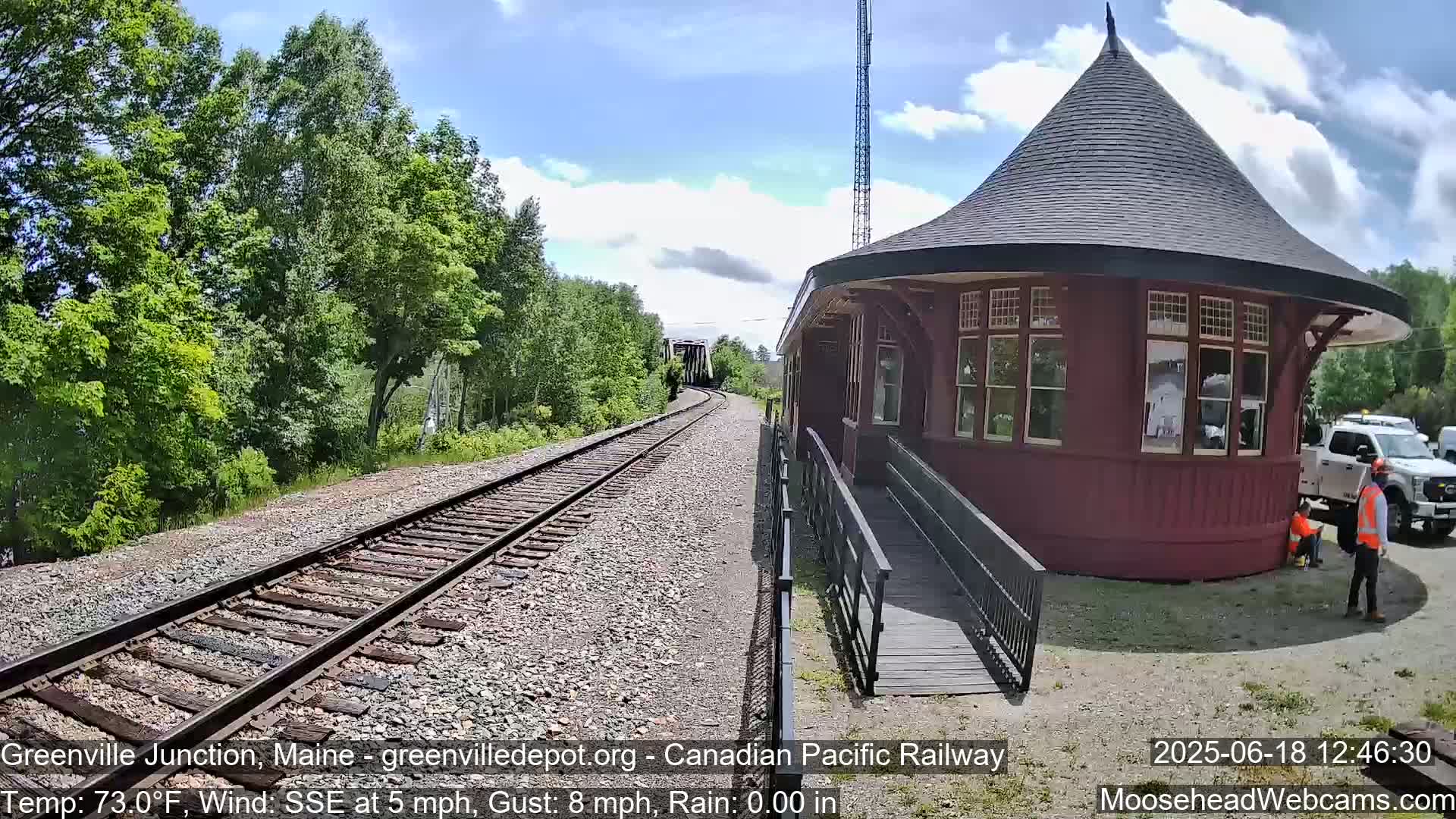 A red, round train depot with a conical roof sits next to railway tracks that curve into a wooded area under a partly sunny sky.