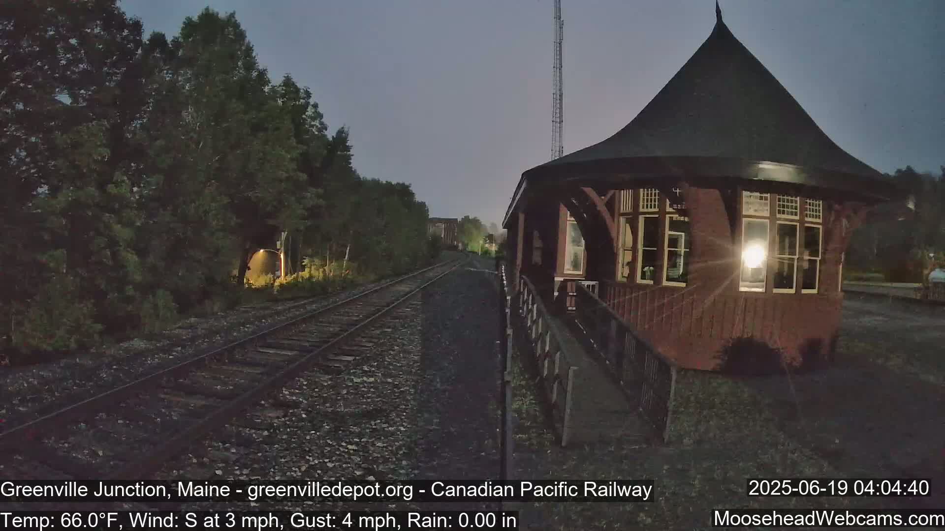 A dark-red train station with a conical roof stands beside railroad tracks at night under a twilight sky, with a slight south wind.