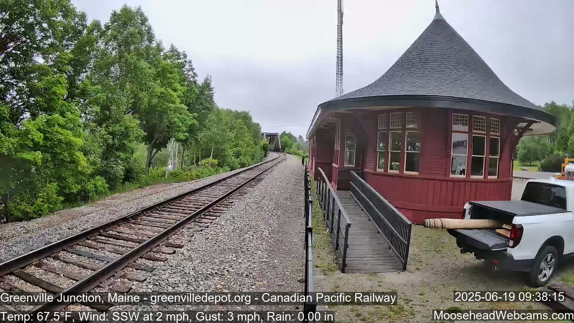 A red, round train station with a ramp sits next to railroad tracks that curve into the distance under a partly cloudy sky, with lush green trees bordering the tracks and a white pickup truck parked nearby.