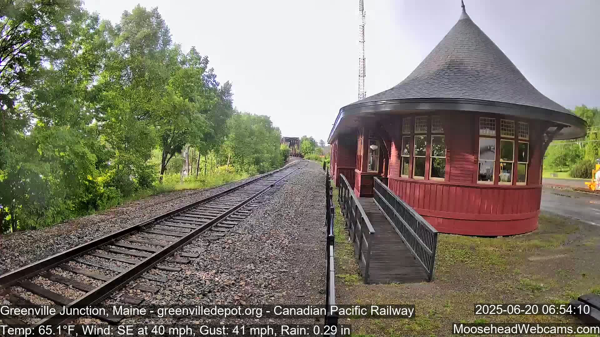 A red, round train station with a conical roof sits beside a railway track in rainy weather, surrounded by green trees.