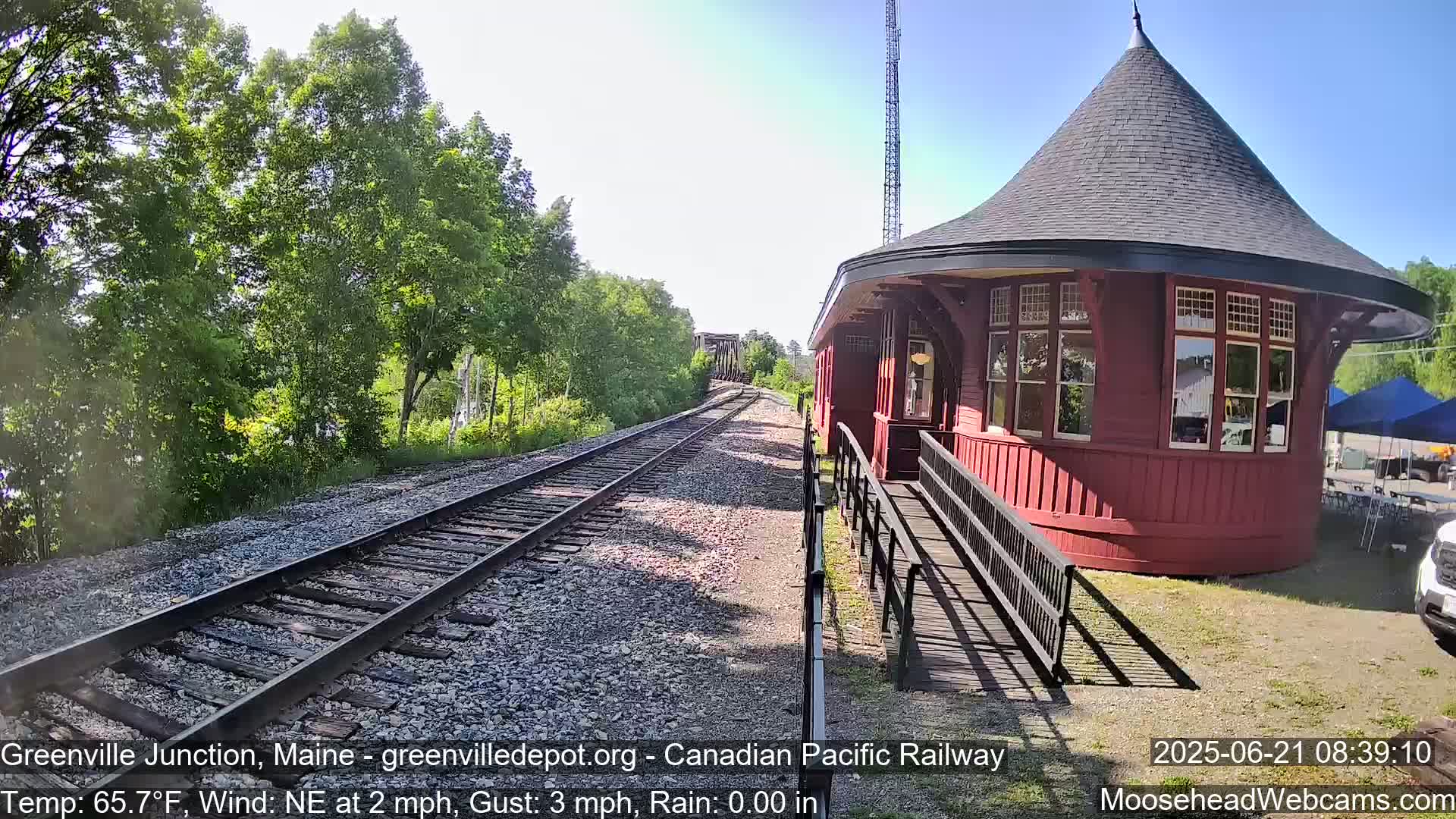 A red, round train station with a conical roof is situated beside railroad tracks on a sunny day with a light northeast wind.