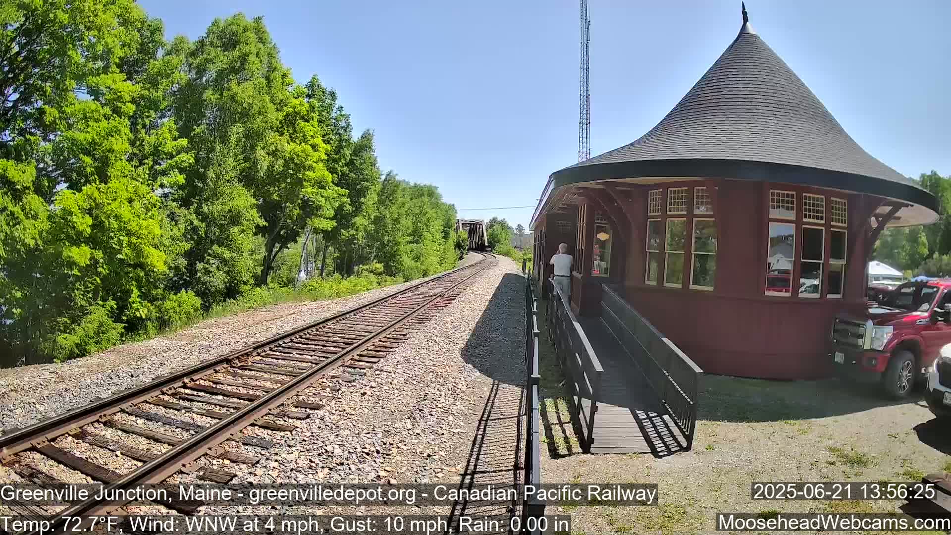 A red train station with a conical roof stands beside railroad tracks on a sunny day with a light wind, and a train is visible in the distance.