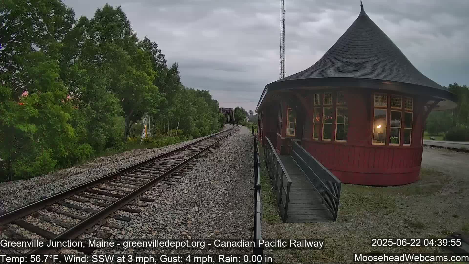 A red, round train depot with a conical roof sits beside railway tracks that curve into a wooded area under a cloudy sky with light rain.