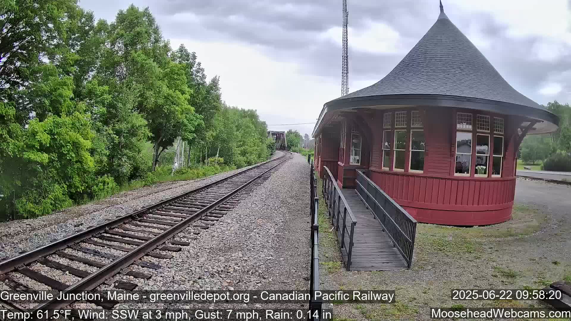 A red, round train depot with a ramp sits beside railroad tracks that curve into a treeline under a rainy, overcast sky.