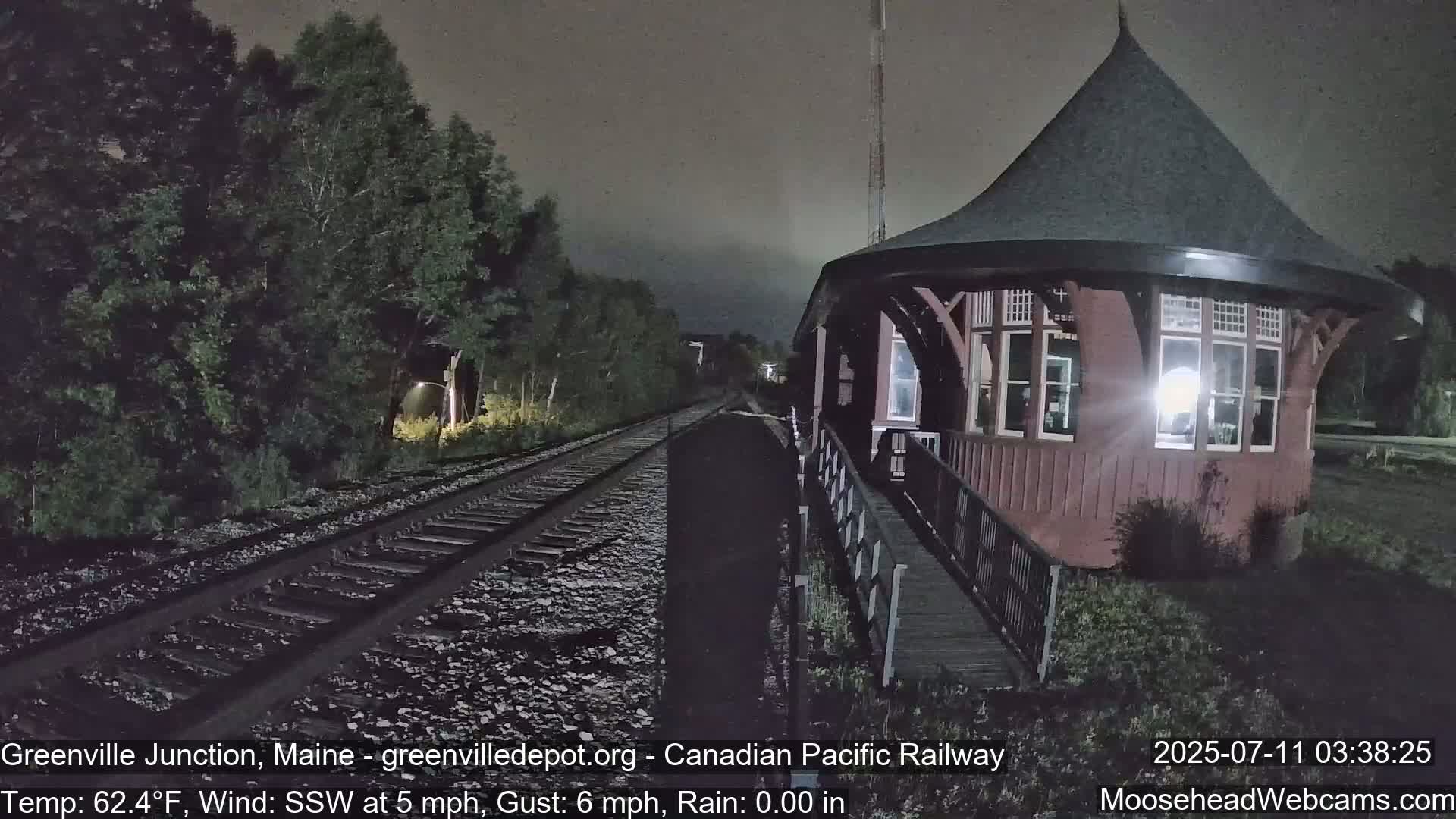 A red train depot with a conical roof stands beside train tracks at night in rainy conditions.
