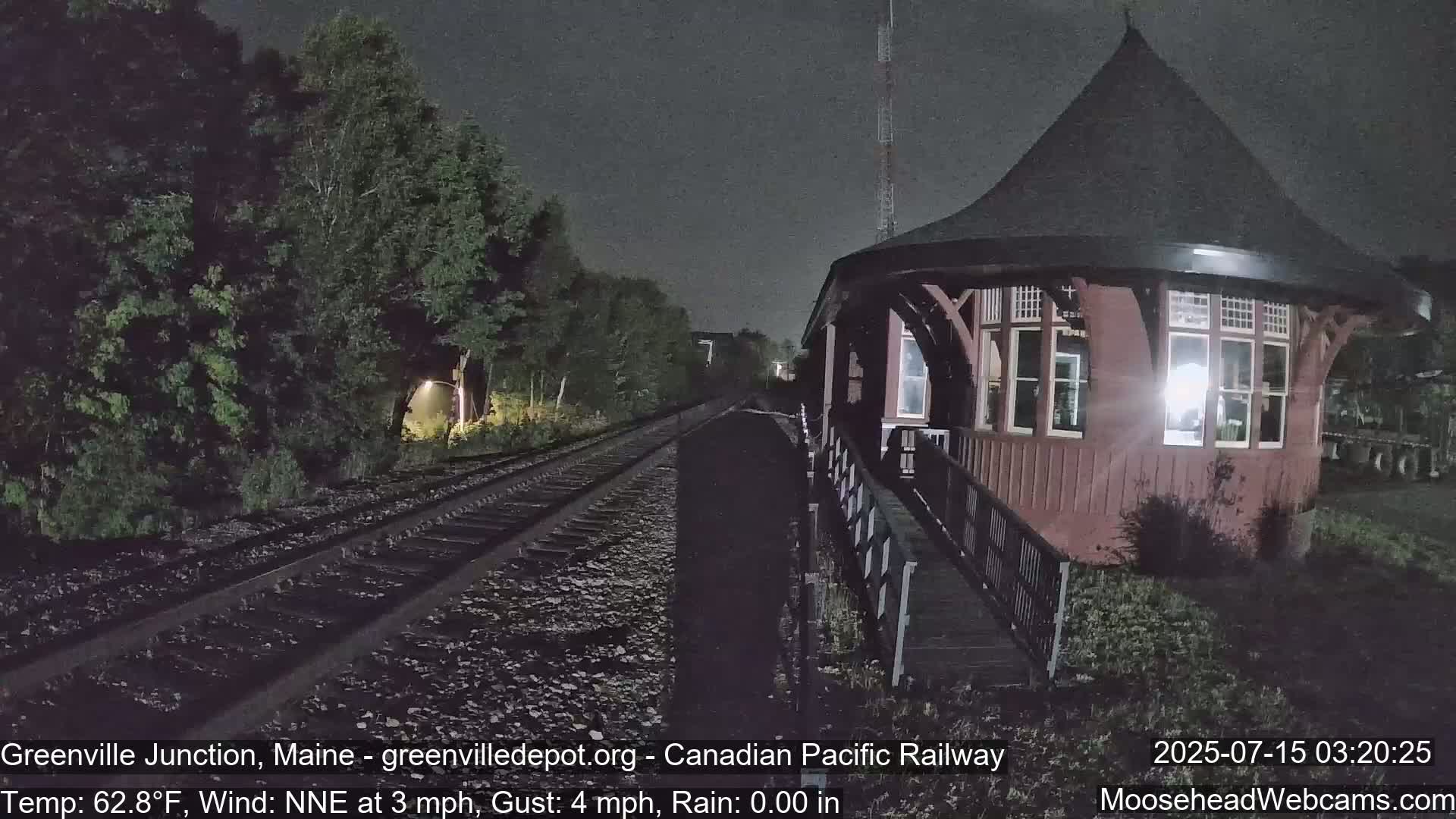 A dark red, octagonal train station with a conical roof stands next to railroad tracks at night under a clear sky, with calm winds.
