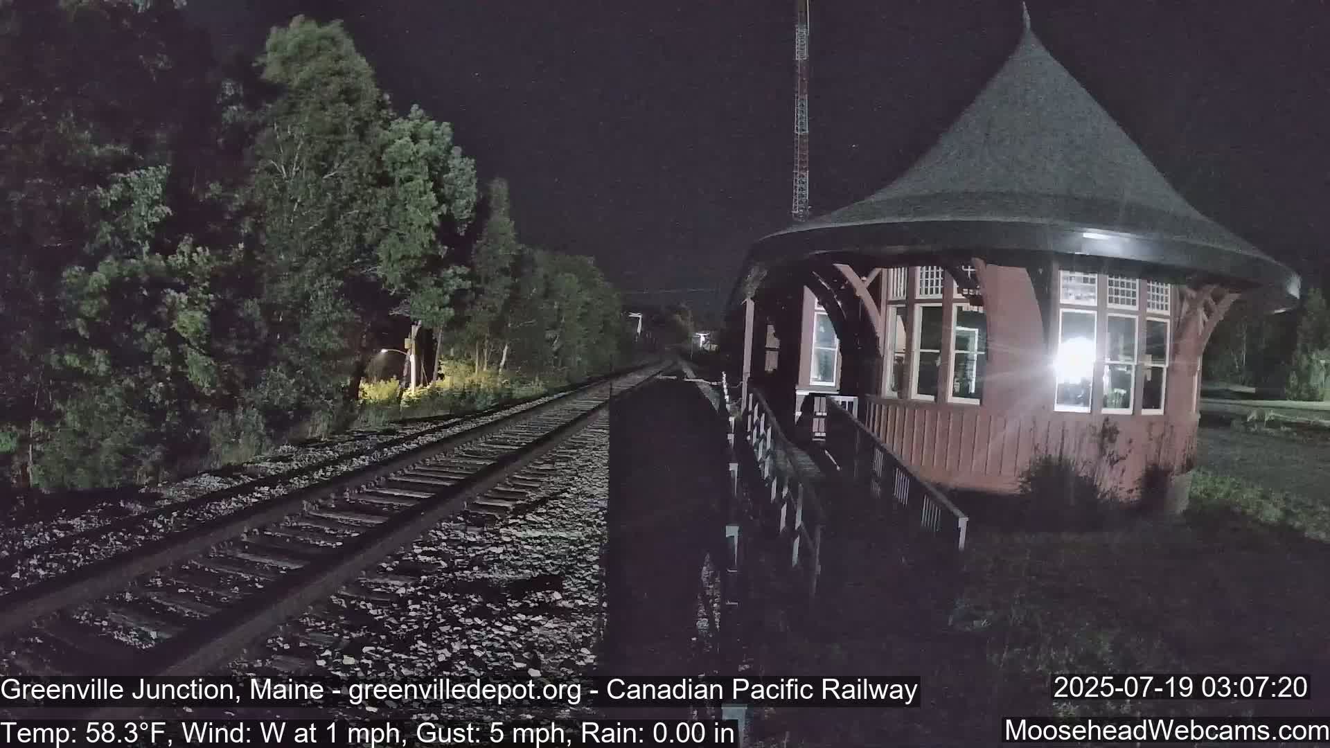 A nighttime view of railroad tracks leading to a round, reddish-brown train depot under a dark sky with a light westerly wind.