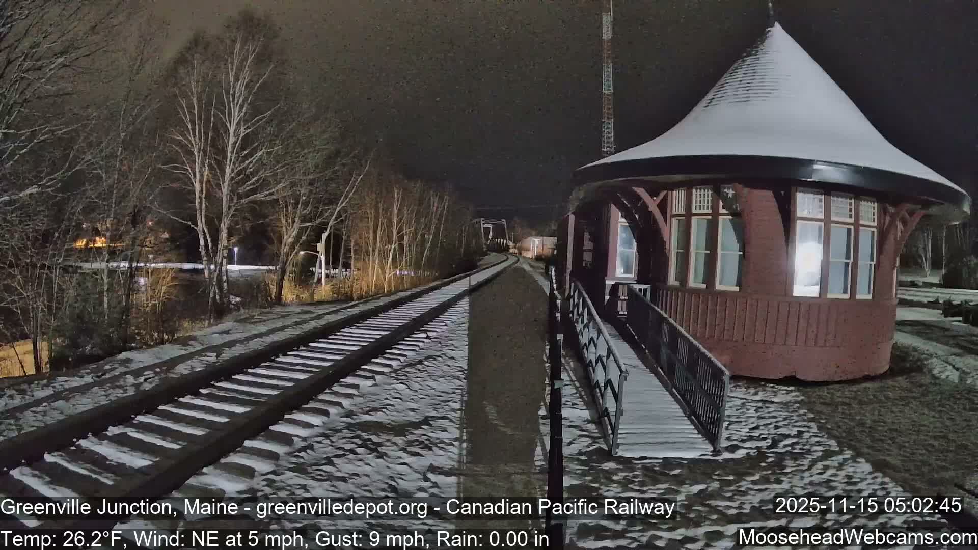 A vintage-style, reddish-brown circular building with a snow-covered conical roof stands beside snow-dusted railroad tracks and bare trees, all illuminated under a dark, clear night sky on a cold winter evening.