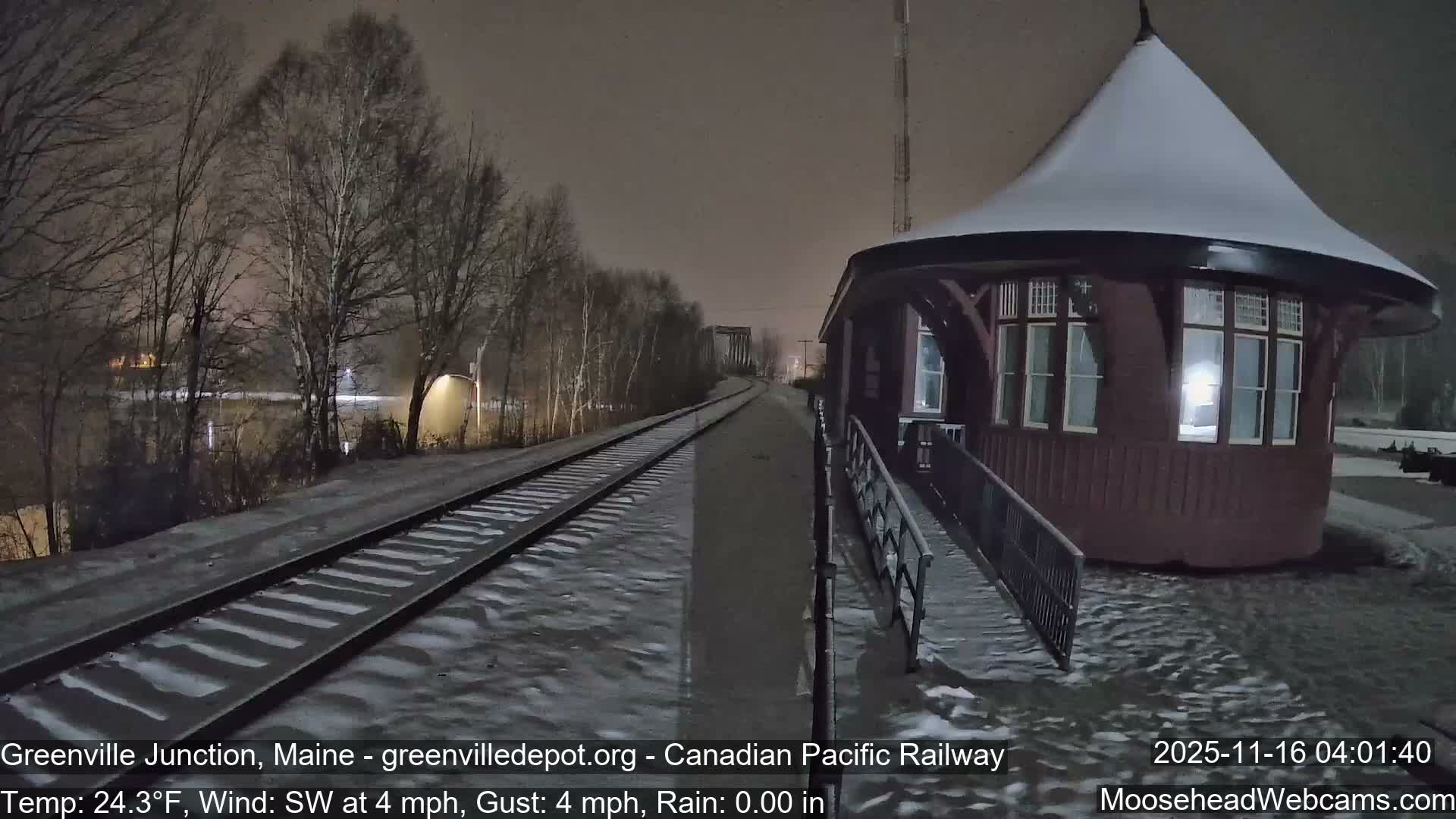 A snow-covered train station with a distinctive conical roof stands beside railway tracks and bare trees overlooking a body of water on a dark and snowy night.
