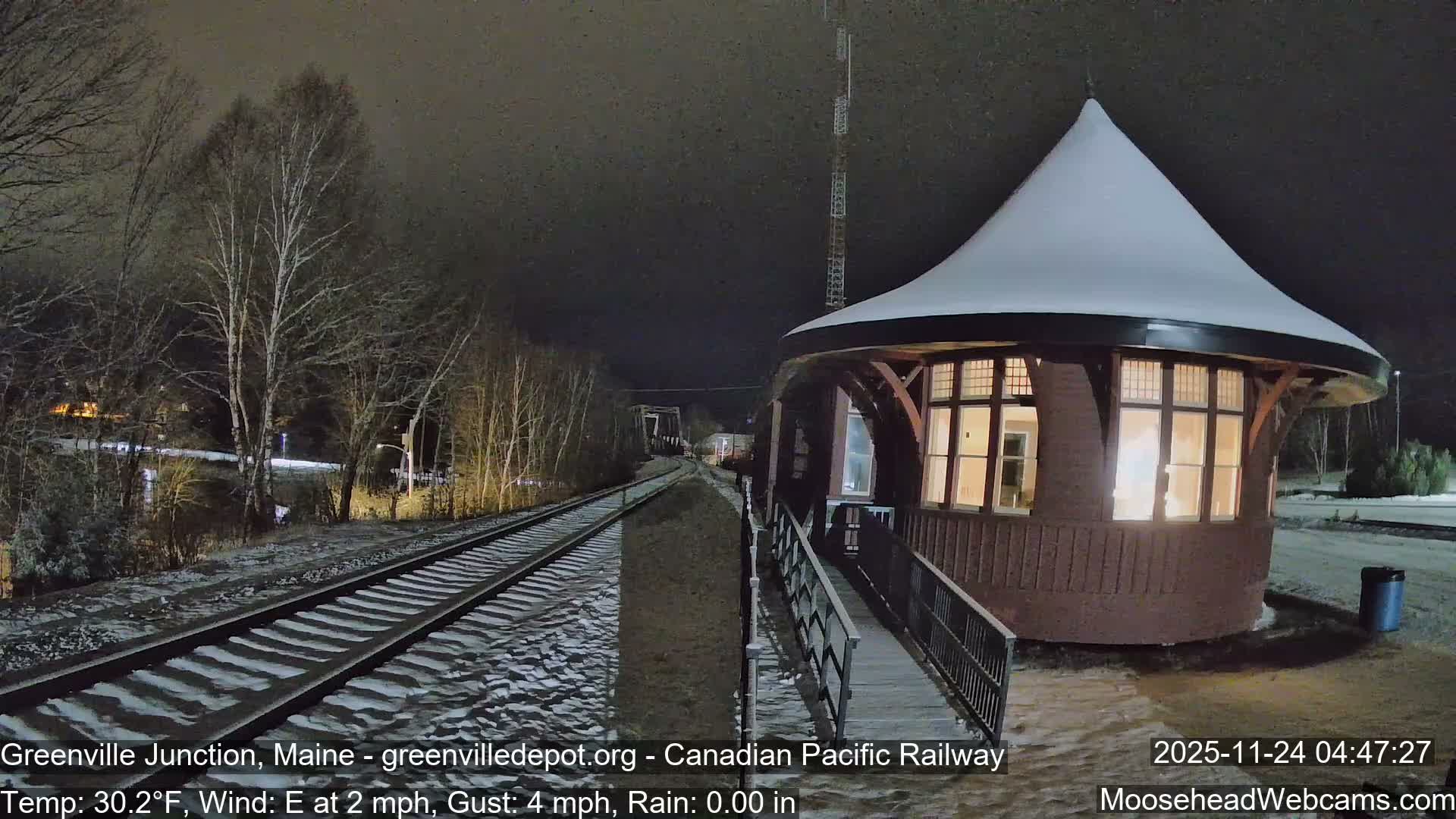At night, a warmly lit, round building with a snow-covered conical roof sits next to snow-dusted railway tracks that lead past bare trees and a distant bridge under a dark, overcast sky.