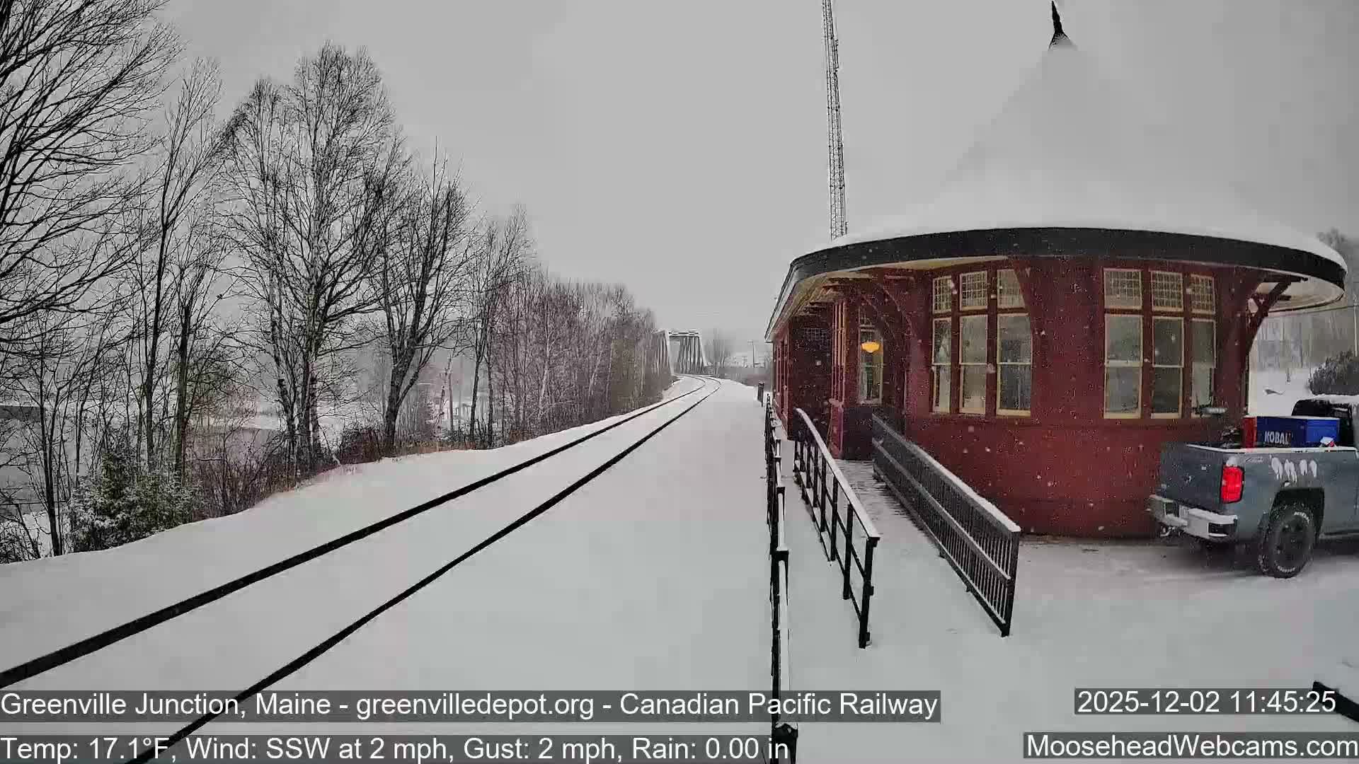 A snow-covered historic train depot with a red rounded building sits beside parallel train tracks, all blanketed in fresh snow under an overcast sky with light snowfall, while bare trees line the distant left bank and a bridge spans further down the tracks.