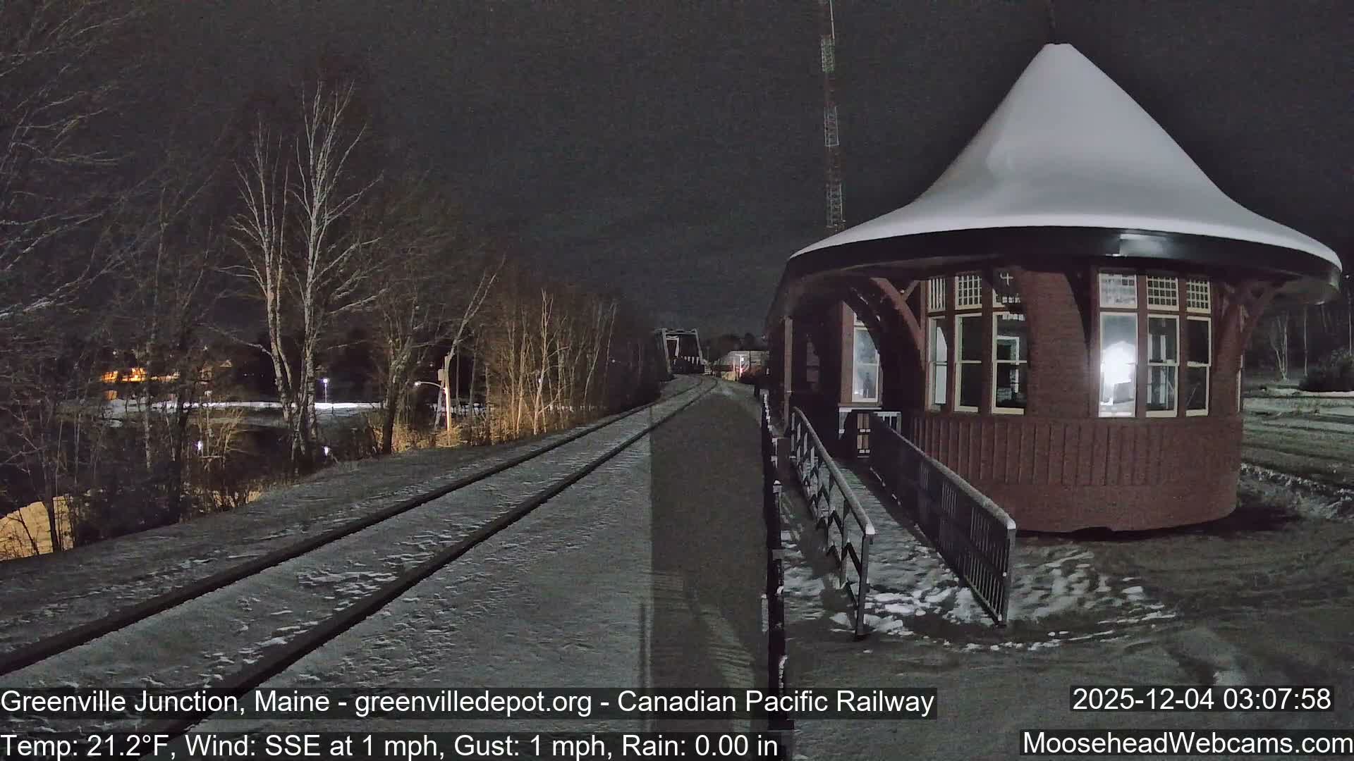 A snow-dusted brick building with a distinctive white conical roof stands beside snowy railroad tracks and bare trees on a clear, cold winter night.