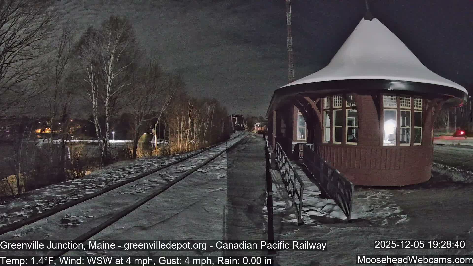 A cold, snowy night scene reveals a conical-roofed train station beside snow-dusted railroad tracks that extend past a body of water and bare trees.