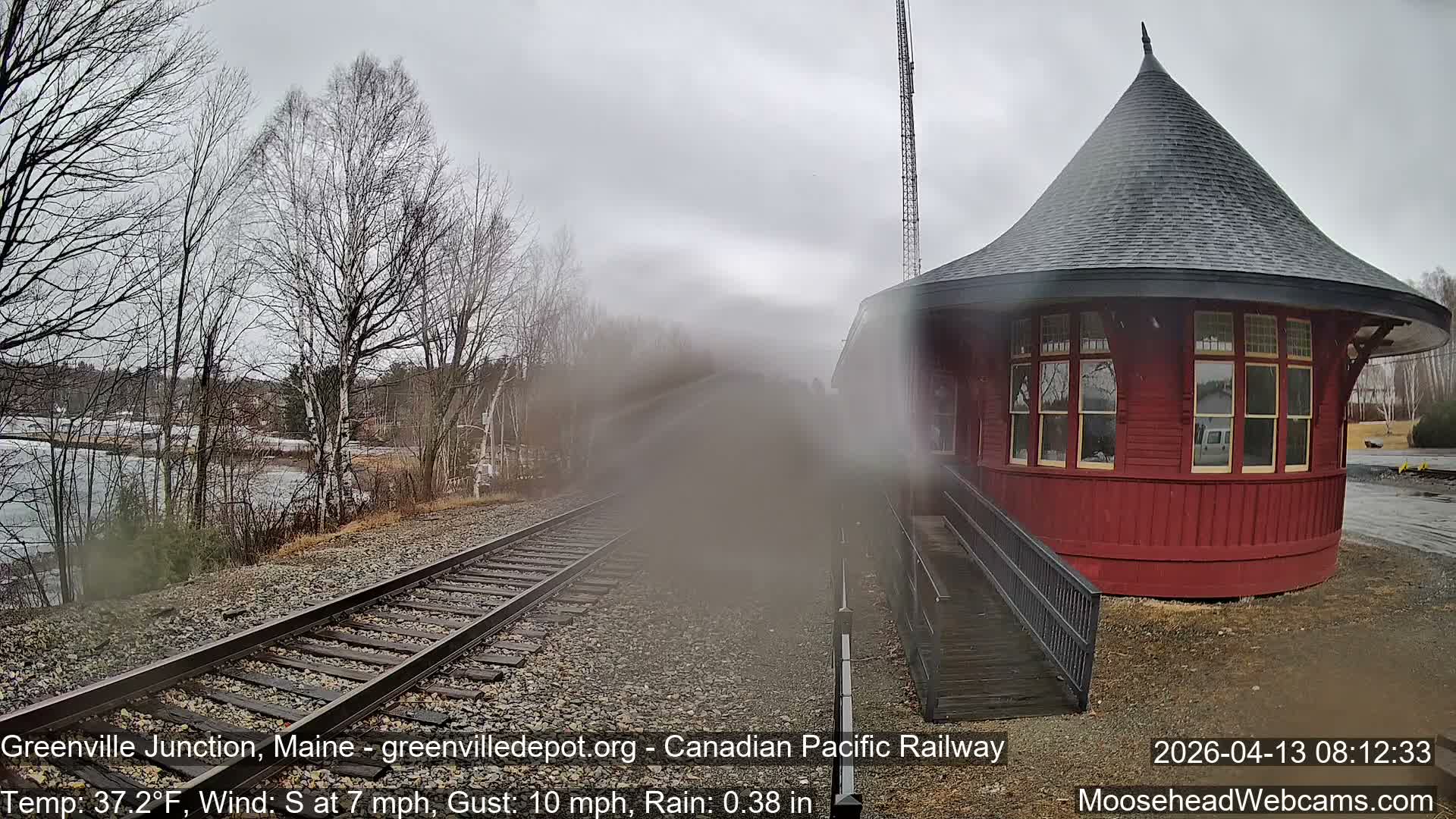 A cold, snowy night scene reveals a conical-roofed train station beside snow-dusted railroad tracks that extend past a body of water and bare trees.