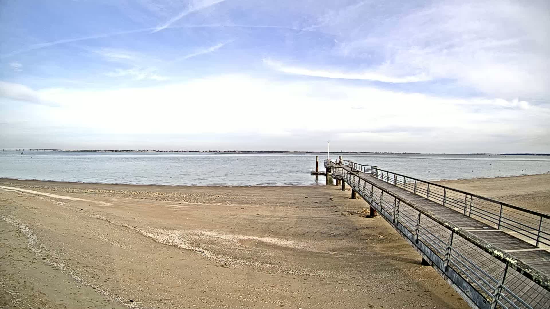 A long pier extends from a sandy beach into a calm body of water under a partly cloudy sky.
