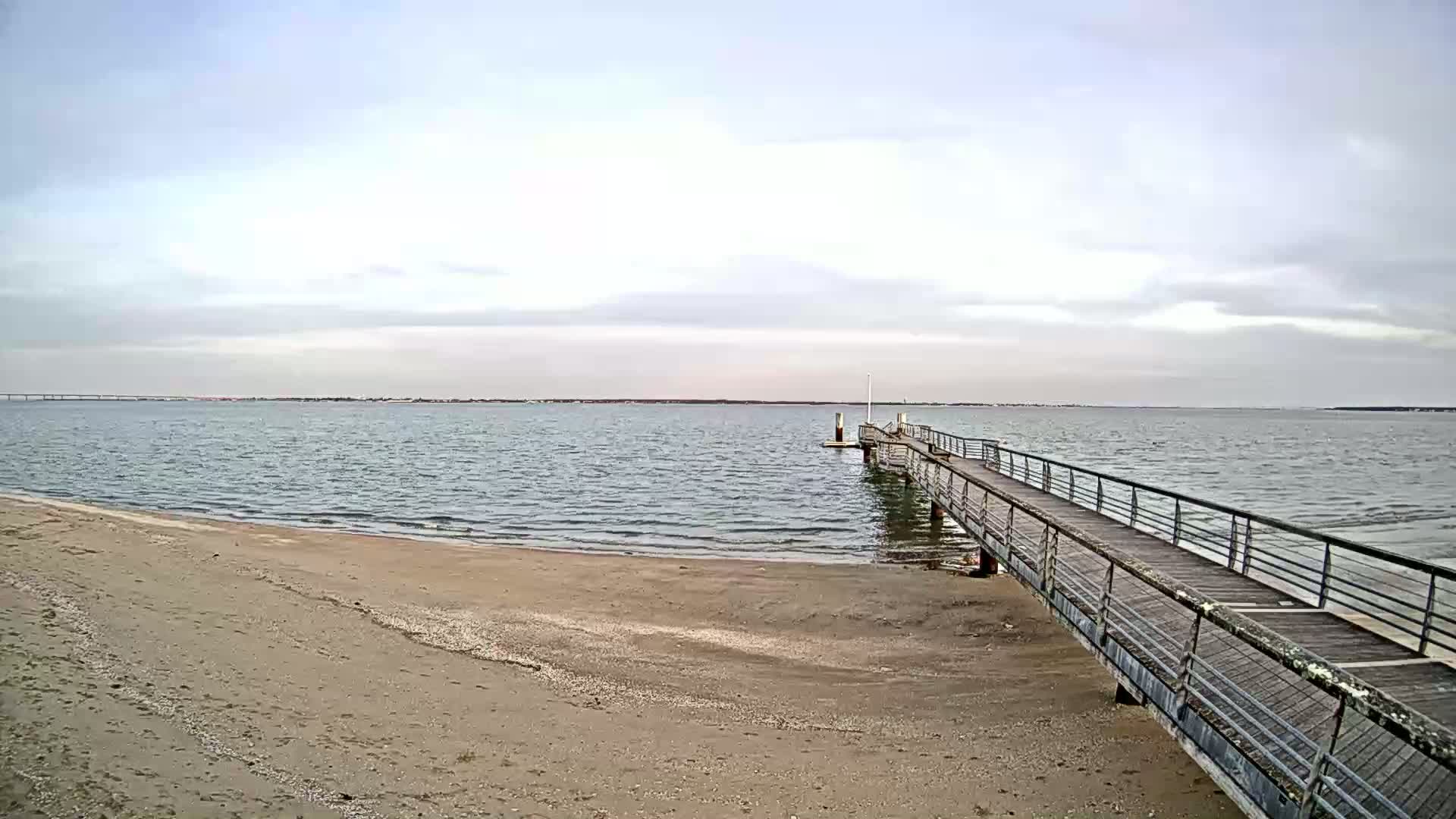 A sandy beach slopes gently down to a calm, gray body of water where a long pier extends out.