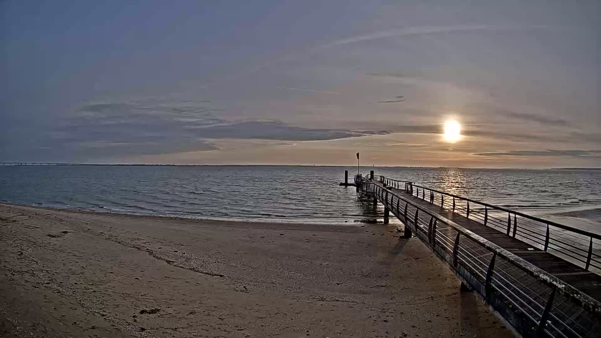 A long pier extends from a sandy beach into calm water under a partly cloudy sunset.
