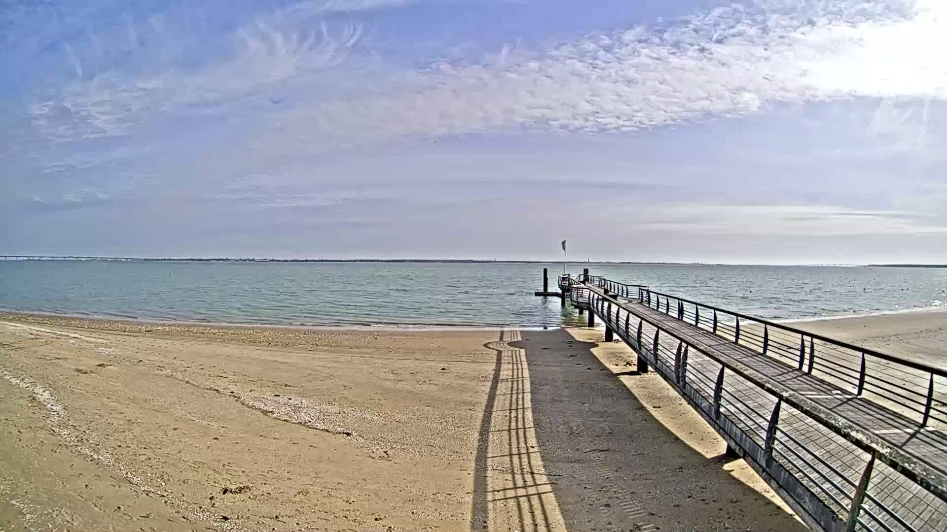 A long pier extends from a sandy beach into a calm body of water under a partly cloudy sky.