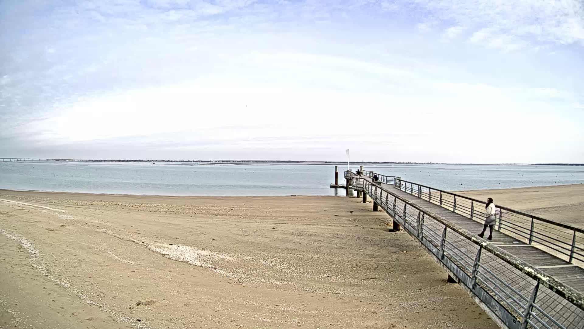 A long pier extends from a sandy beach into a calm body of water under a mostly cloudy sky.