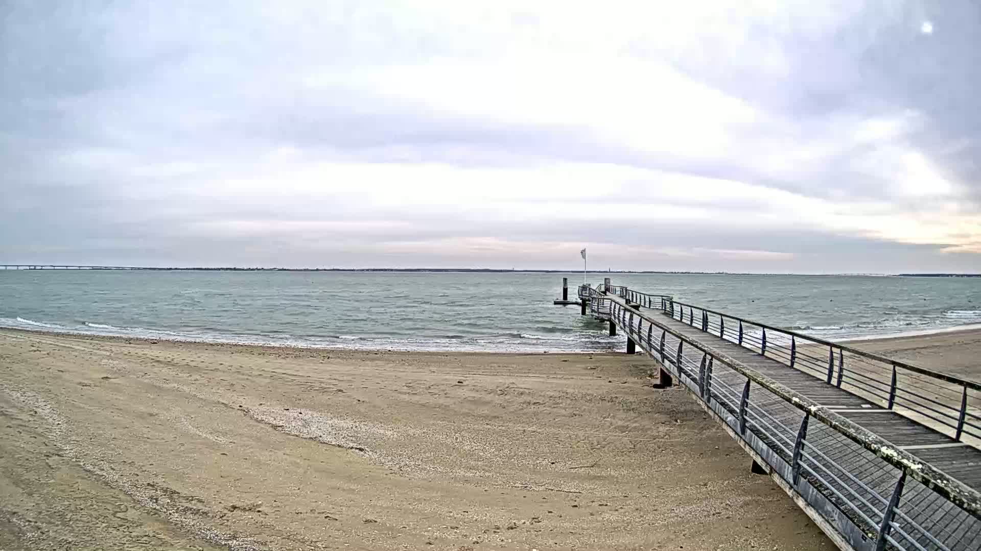 A long wooden pier extends from a sandy beach into a calm, gray sea under an overcast sky.