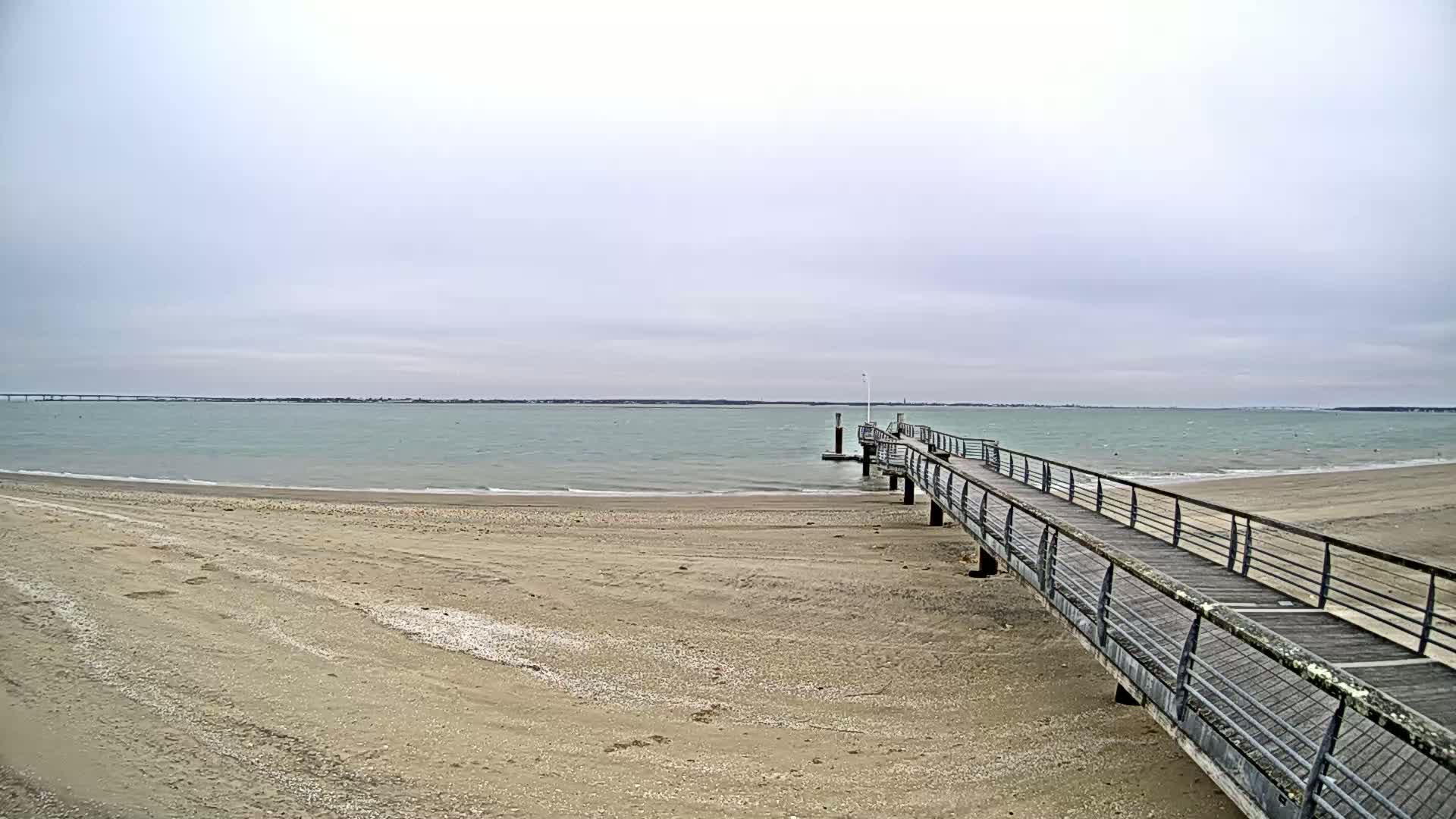 A long pier extends from a sandy beach into a calm, light-green sea under an overcast sky.