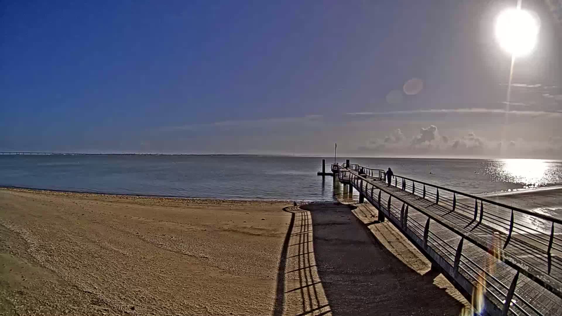 A long pier extends from a sandy beach into a calm, sunlit ocean under a mostly clear blue sky.