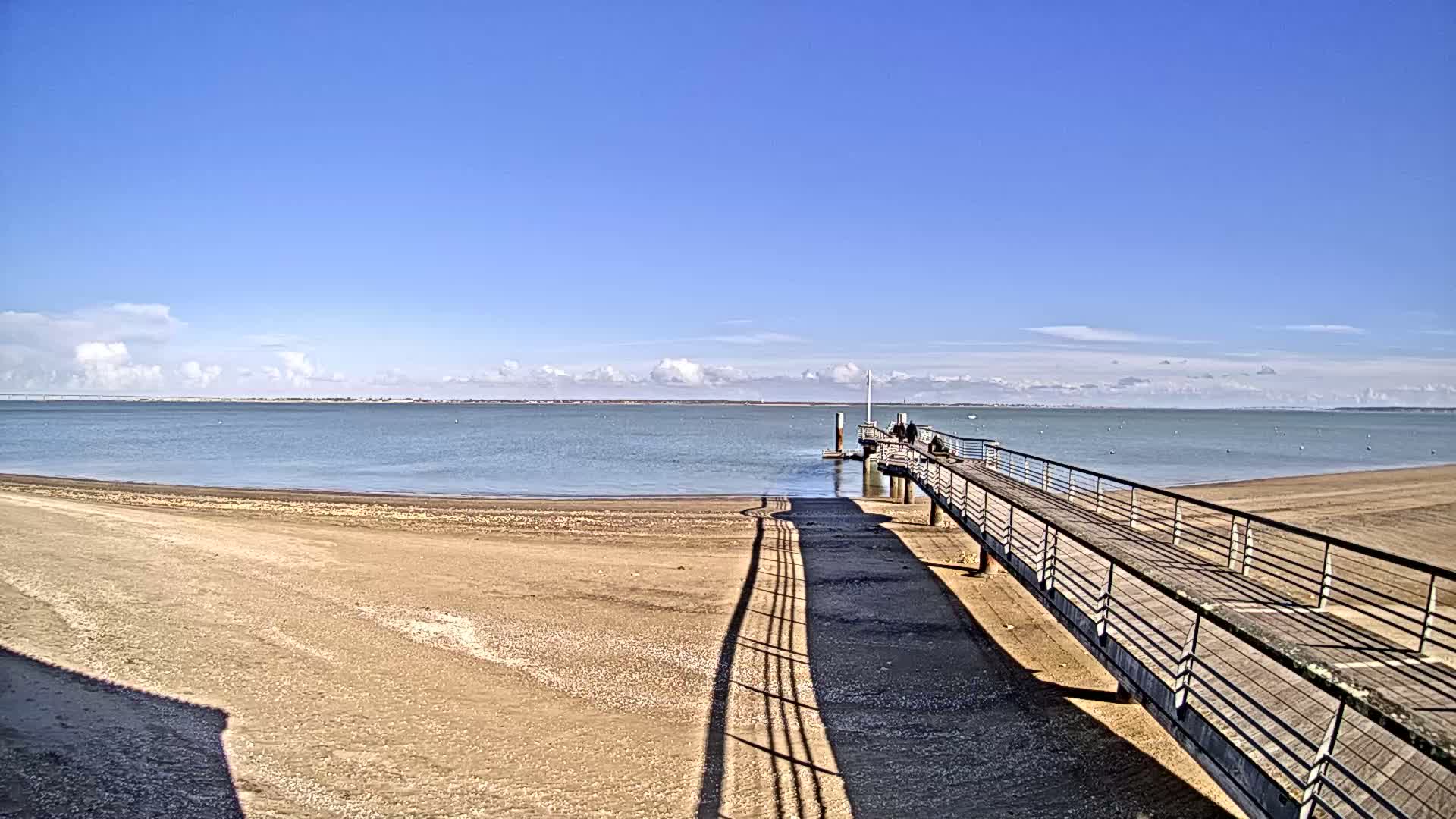 A long pier extends from a sandy beach into a calm, blue body of water under a mostly clear, sunny sky.