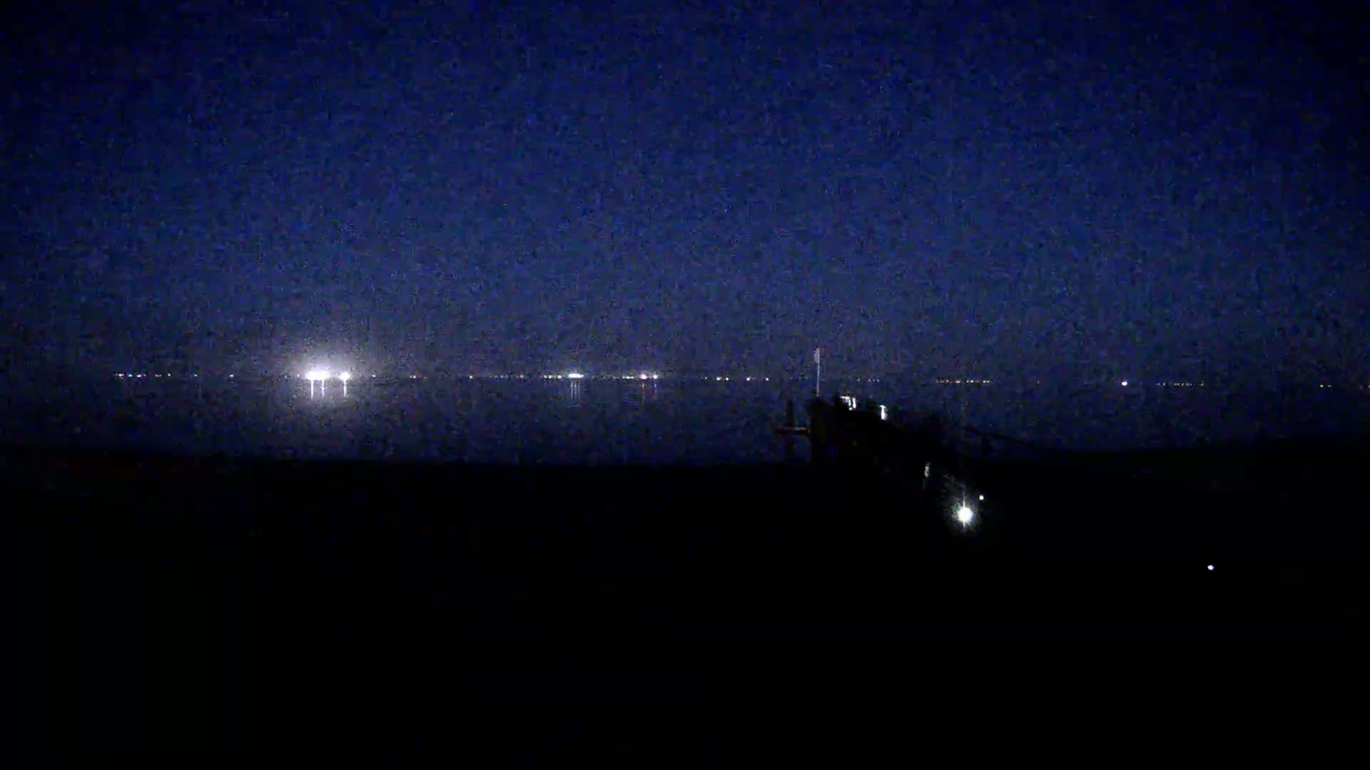 Under a dark night sky, a body of water reflects distant lights, while a dark structure sits in the foreground.