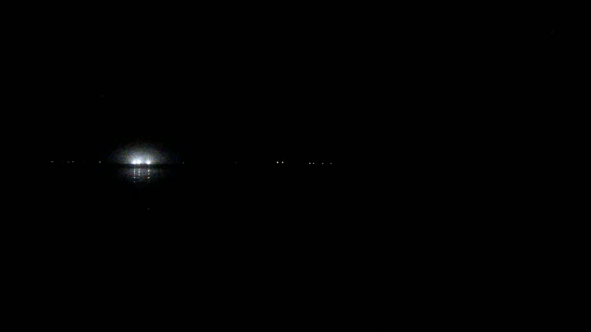 A bright light on a body of water is visible at night, with a few smaller lights faintly visible in the distance.