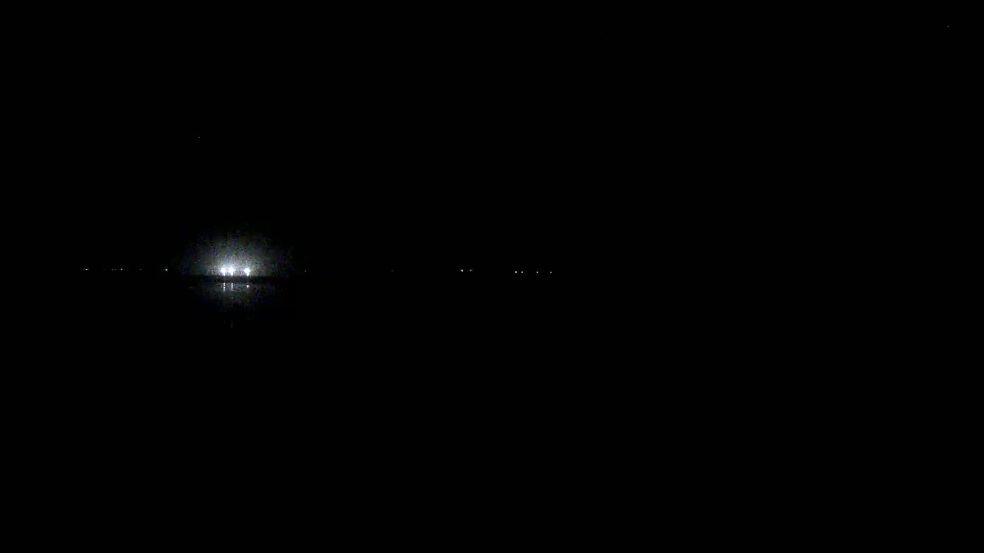 A bright light source is visible on a dark body of water at night, with several dimmer lights faintly visible in the distance.