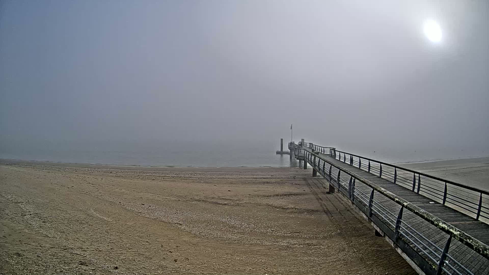 A long pier extends from a sandy beach into a foggy sea under a hazy sun.